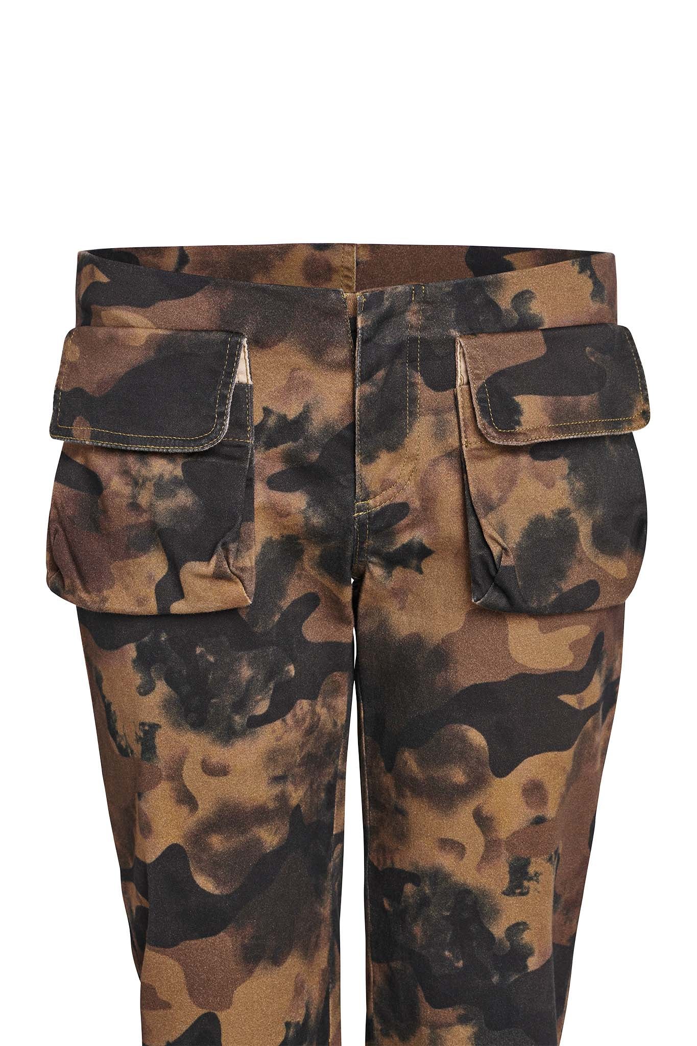 The KIMANI PANT - GREEN : CAMO from I.AM.GIA, featuring shades of brown, black, and green, includes two large pockets with flaps on the sides near the hips. The pants are displayed from the waist down, showcasing their intricate camouflage pattern and stitching details.