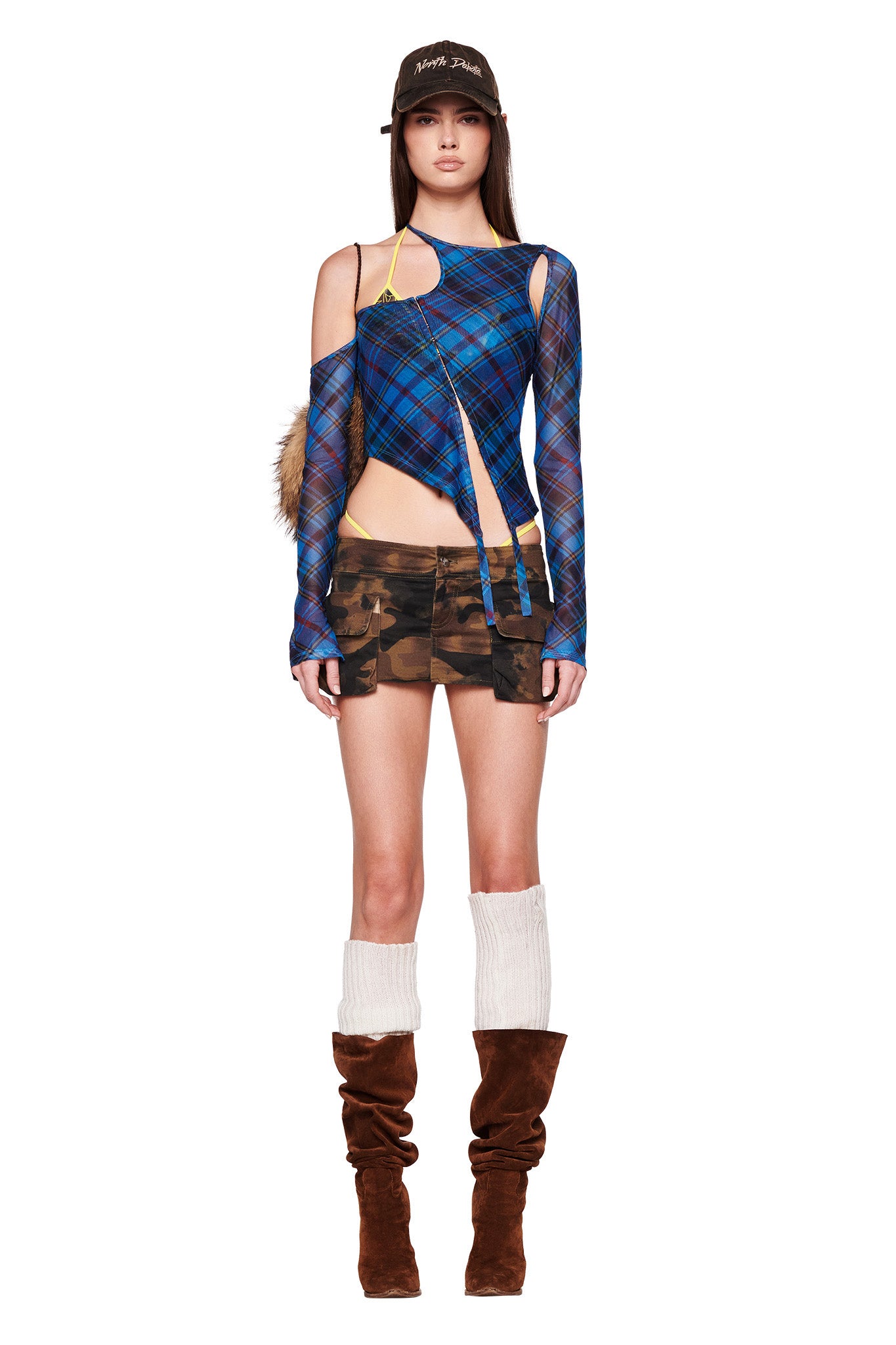A person stands against a white background, wearing the I.AM.GIA ANAFER TOP - BLUE PLAID with cutouts, a short camo-patterned skirt, and brown slouched knee-high boots with white leg warmers. They also have a brown baseball cap on with the words "Paris, Texas" printed on it.
