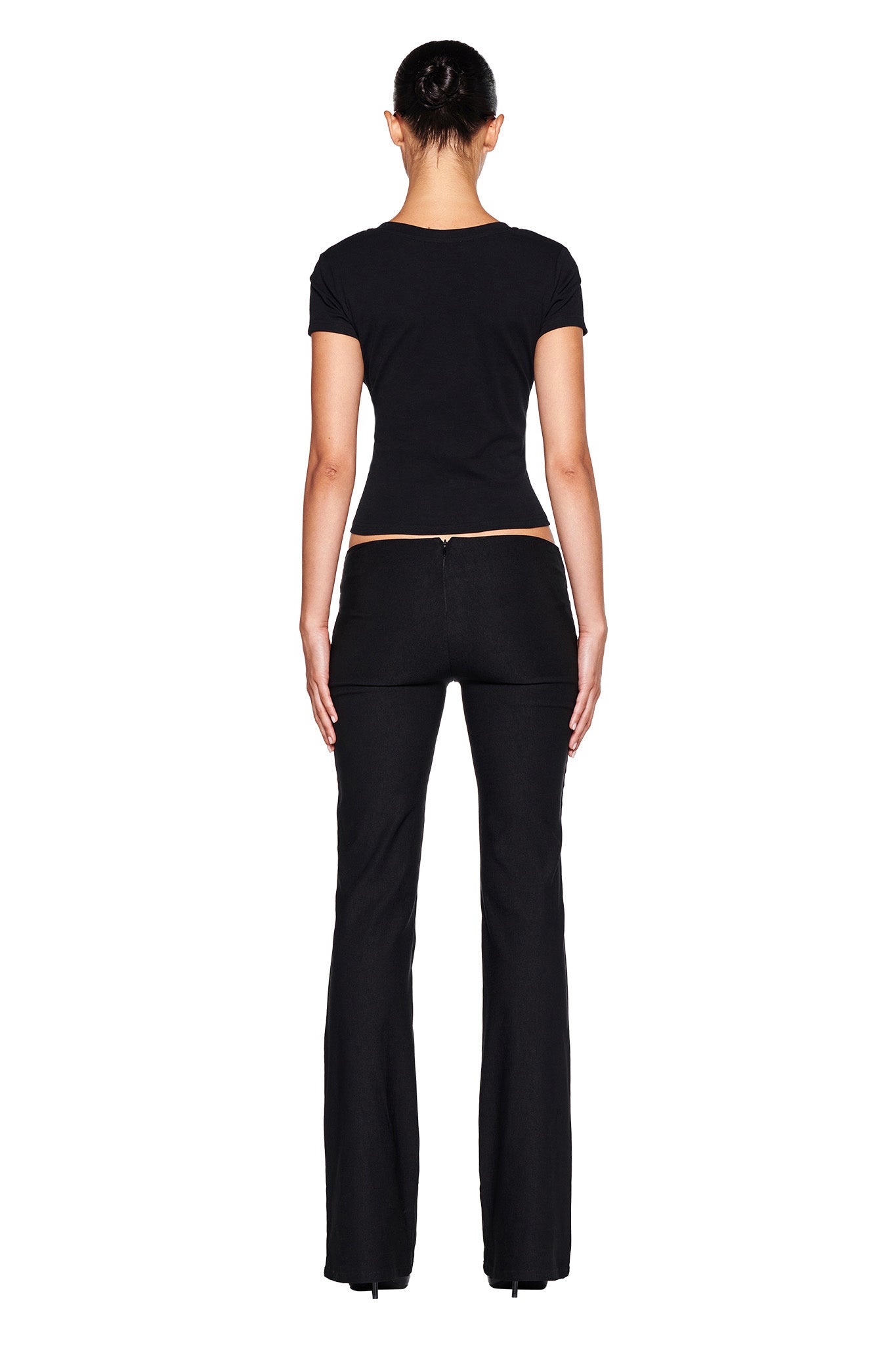 A woman is standing with her back to the camera, showcasing the I.AM.GIA SHARAH PANT in black—a fitted belted suit pant with a flared leg. She pairs it with a sleek black T-shirt and completes her look with a neat bun hairstyle, all set against a plain white backdrop.