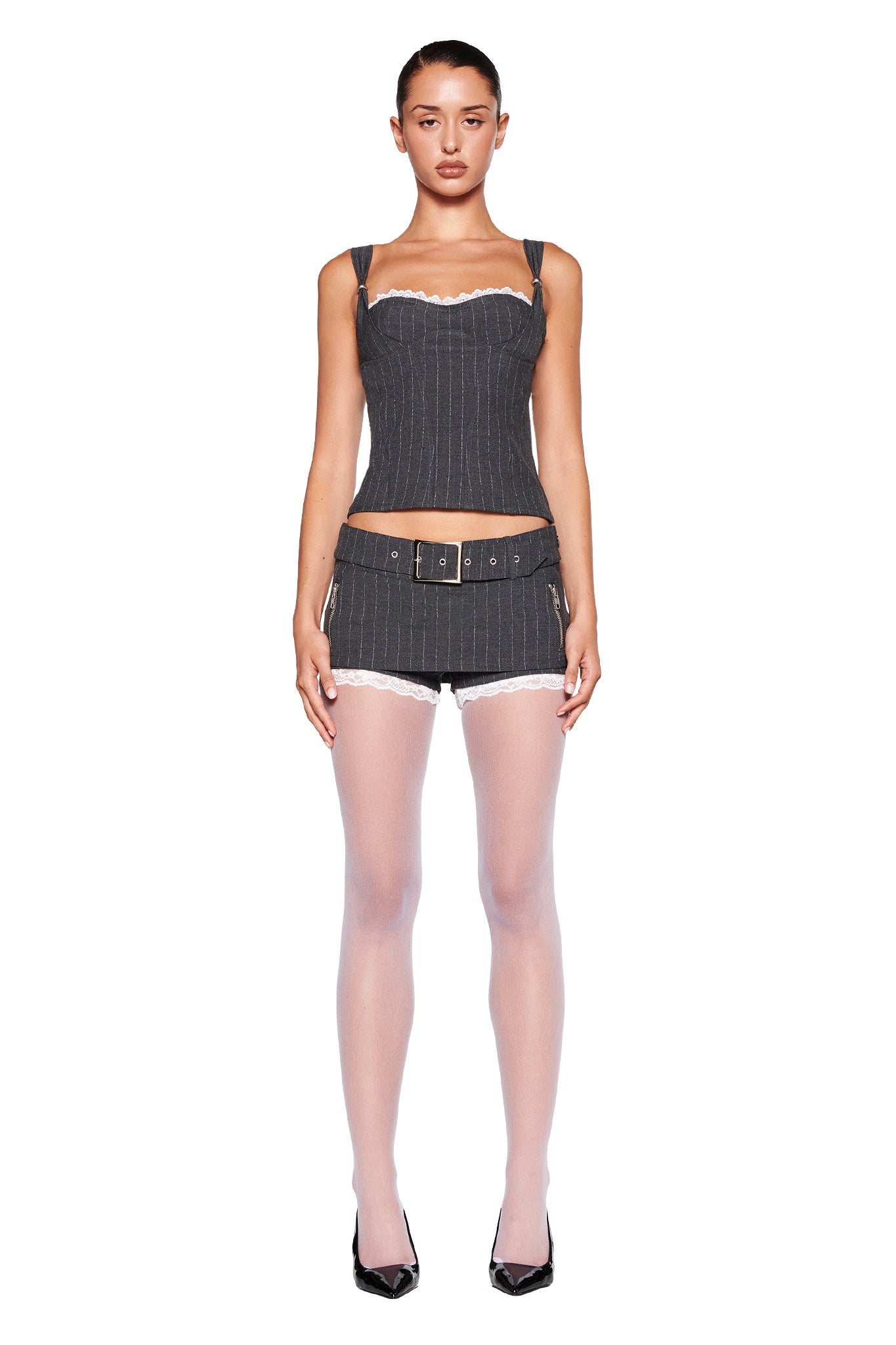 A woman stands against a white background wearing the MARRION CORSET - GREY : PINSTRIPE by I.AM.GIA, paired with a matching tailored mini skirt featuring a belt. She completes her outfit with sheer white tights and black high heels. She has a serious expression and her hair is slicked back.