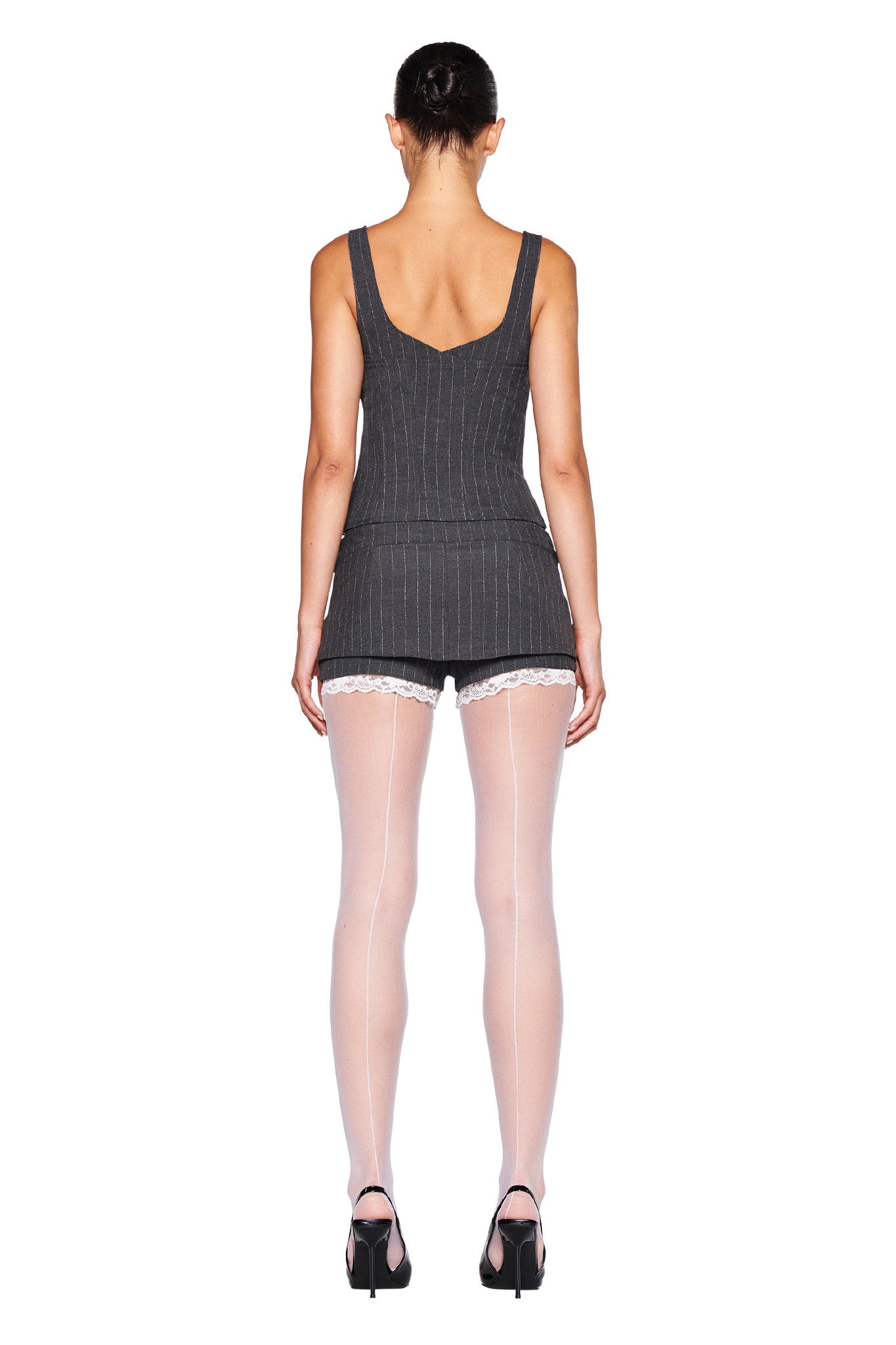 A person with their back to the camera is wearing the MARRION CORSET - GREY : PINSTRIPE from I.AM.GIA, matching pinstriped shorts, white scalloped lace trim underneath the tailored fit shorts, white stockings, and black high-heeled shoes. They have dark hair styled in a bun.