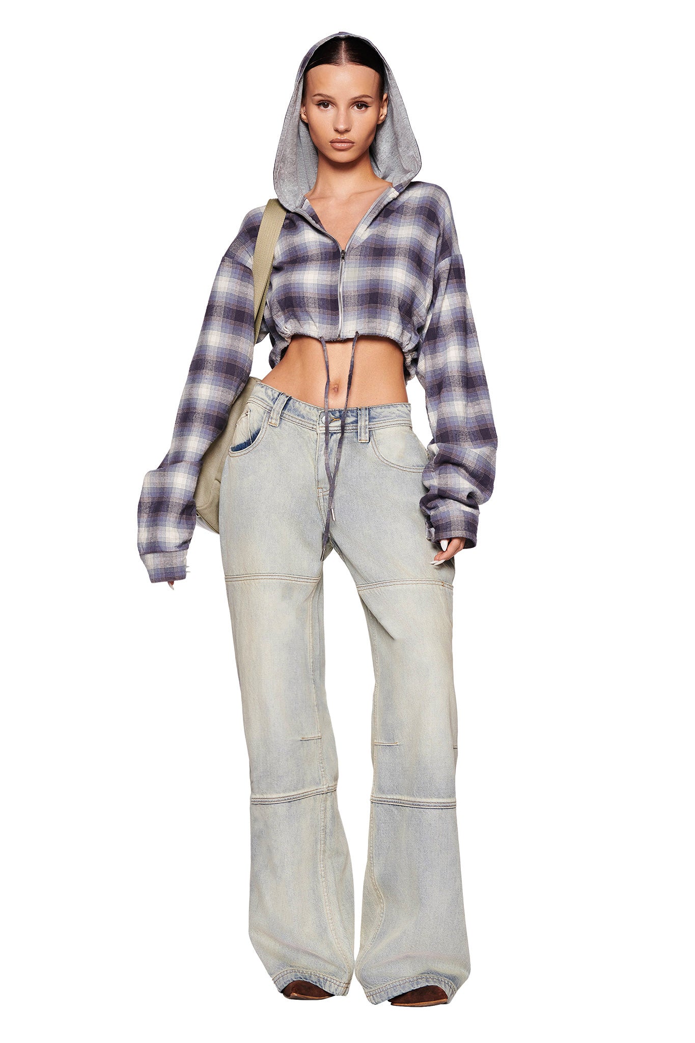 A person stands facing forward, wearing the TIFF JACKET - BLUE, a cropped, long-sleeve flannel shirt with a hood from I.AM.GIA, along with loose-fitting, light-wash jeans. They have a serious expression and are carrying a bag over one shoulder. The background is plain white.