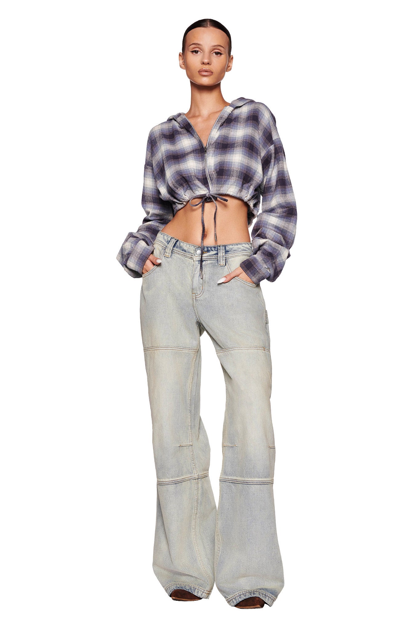 A person poses confidently with hands in pockets, wearing a cropped, long-sleeve TIFF JACKET - BLUE from I.AM.GIA, featuring a plaid pattern in shades of purple, blue, and white. They have paired it with light blue, high-waisted, wide-leg jeans. The plain white backdrop highlights the chic outfit.