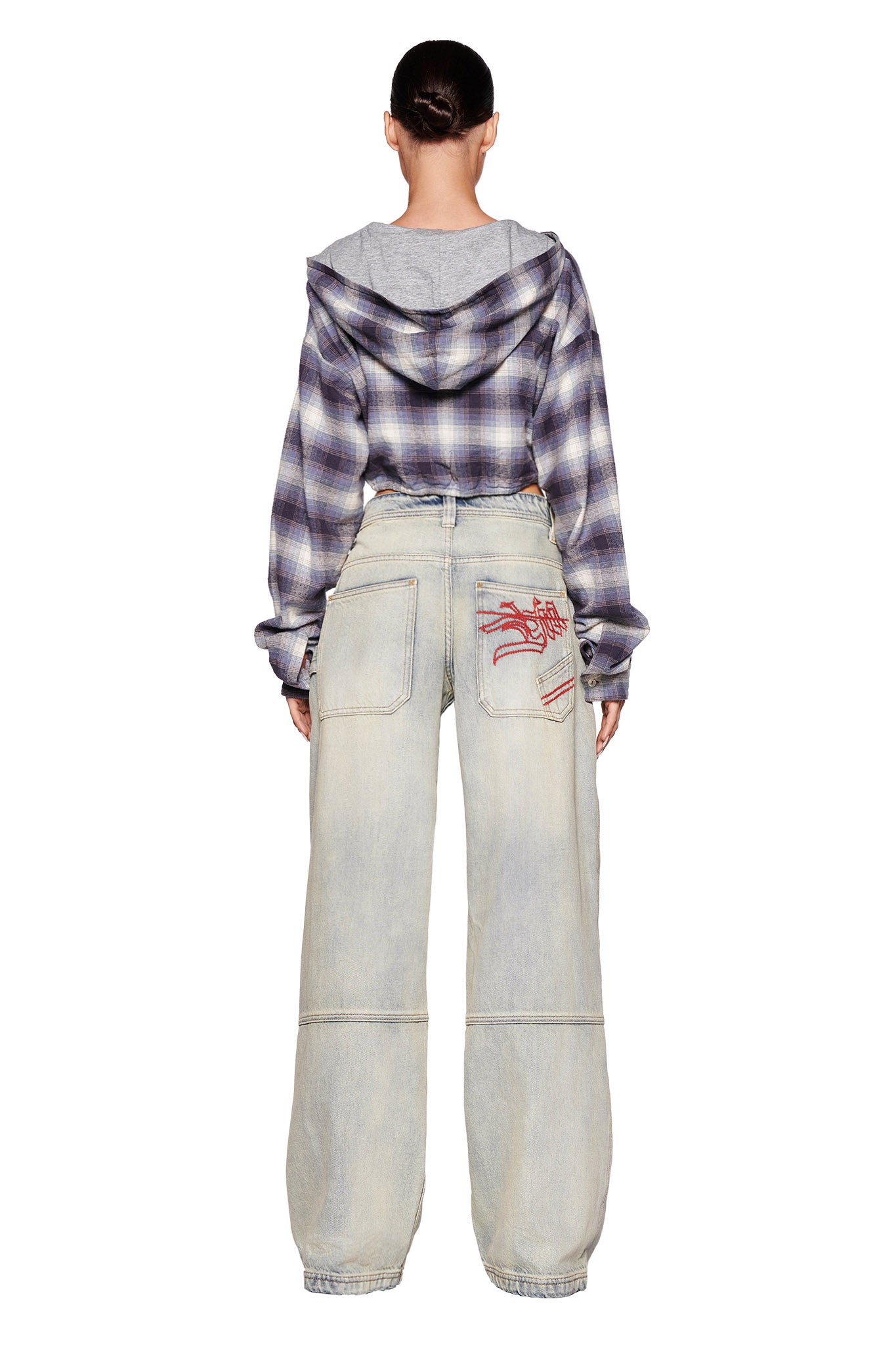 A person is standing with their back to the camera, wearing a purple plaid hooded shirt and baggy, light-wash jeans from I.AM.GIA with large pockets. The left pocket features red abstract embroidery. Their hair is tied back neatly.