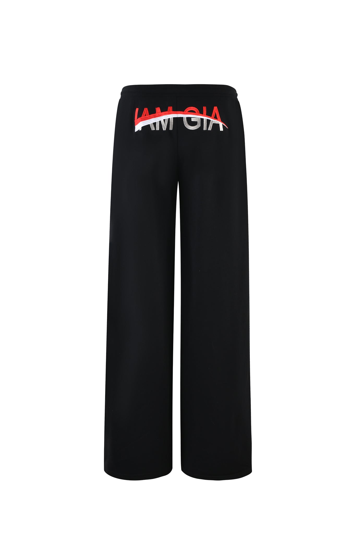 The JUSTINE PANT - BLACK from I.AM.GIA is displayed from the back, featuring a low-rise waistband with "AMGIA" printed in bold white lettering and a red strike-through line. The black baggy track pants offer a relaxed fit without any visible pockets or additional details.