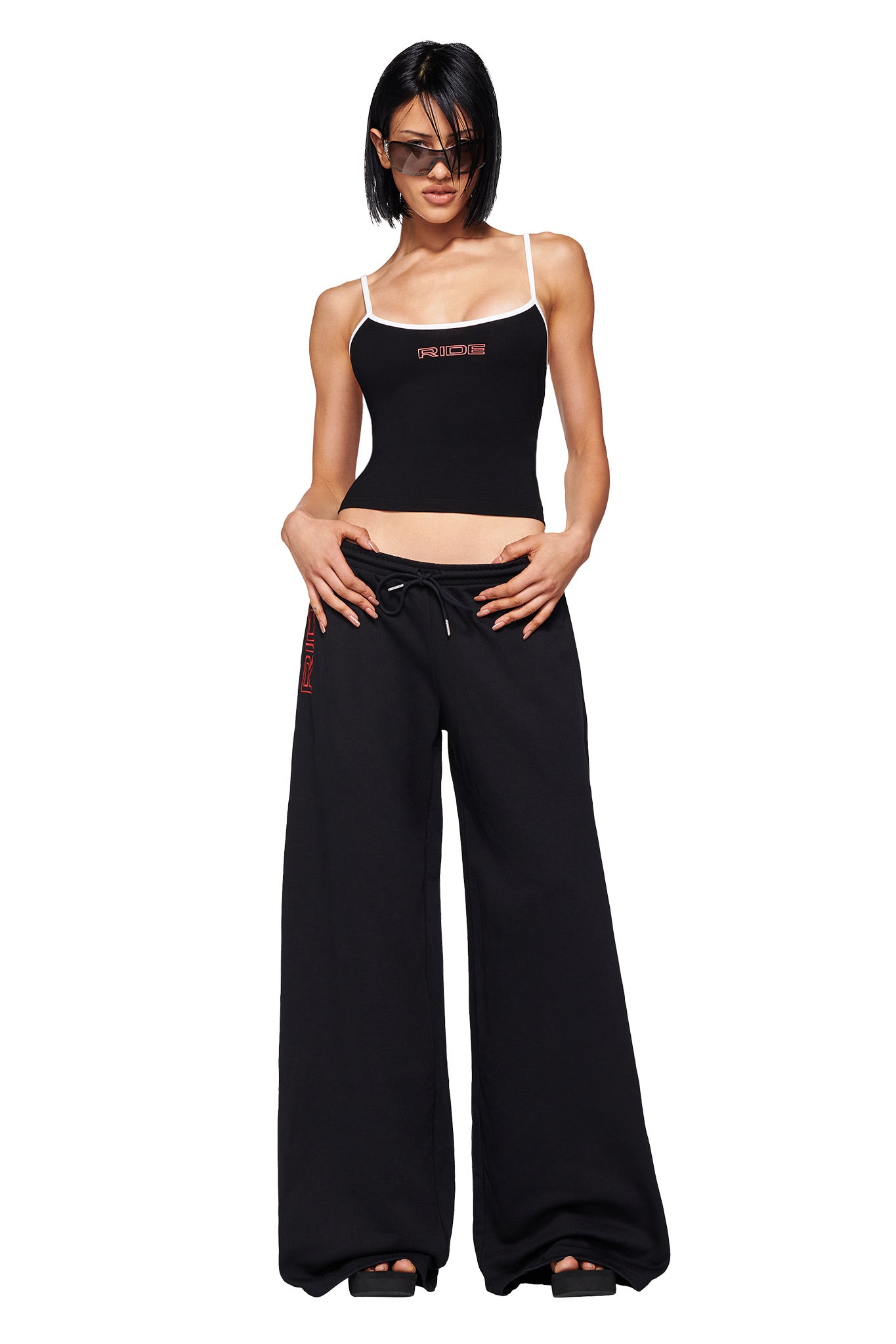 A woman wearing sunglasses, the SHAYLA TANK - BLACK by I.AM.GIA with white straps and graphic lettering, and loose black drawstring pants stands against a white background.