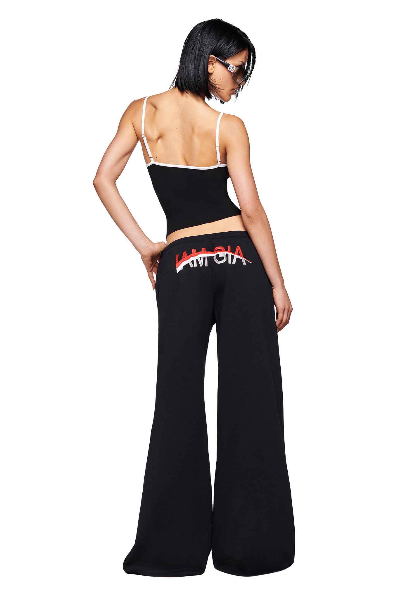 A woman, facing away, wears the I.AM.GIA SHAYLA TANK in black and matching black wide-leg pants with logo waistband, paired with sunglasses. She poses confidently against a plain white background.