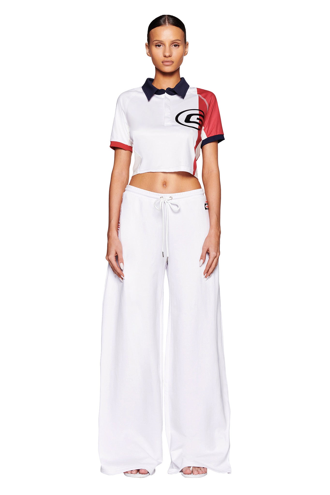 A person stands facing forward, wearing a cropped short-sleeve polo shirt with a red, white, and blue color scheme and a large emblem on the chest. They are also wearing high-waisted, wide-legged JUSTINE PANTS - WHITE from I.AM.GIA with a drawstring waist. The background is white.