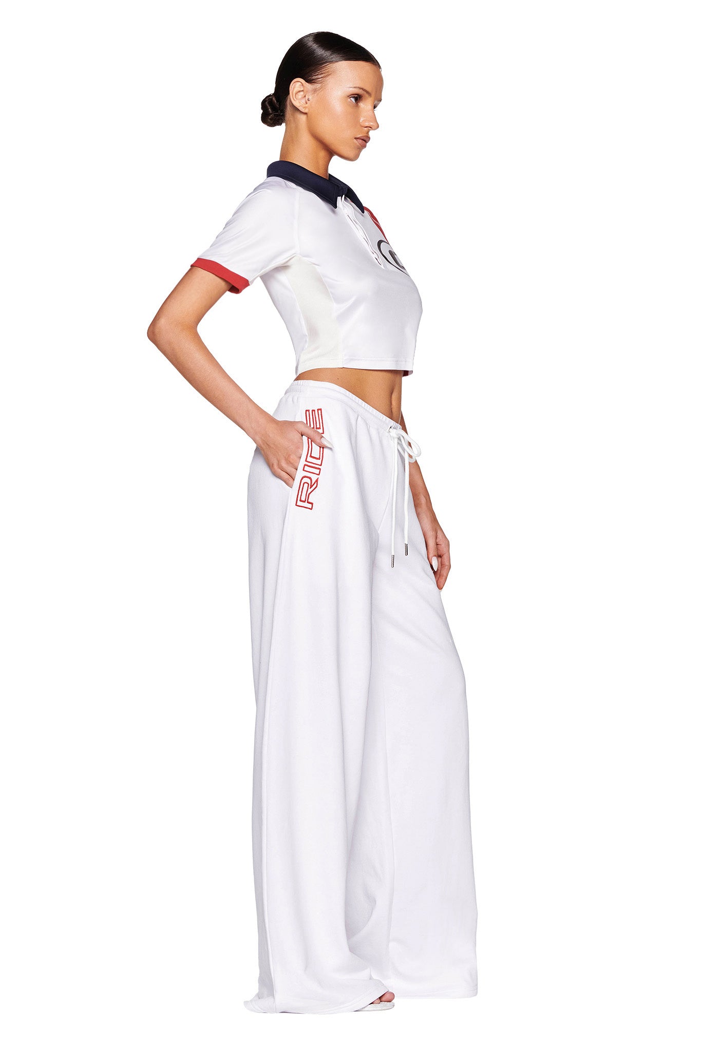 A person is standing in profile against a white background, wearing an I.AM.GIA white cropped polo shirt with red and blue accents. They are paired with the I.AM.GIA JUSTINE PANT - WHITE, which features a loose fit and "RUE" printed near the pocket. Their hair is neatly tied back.