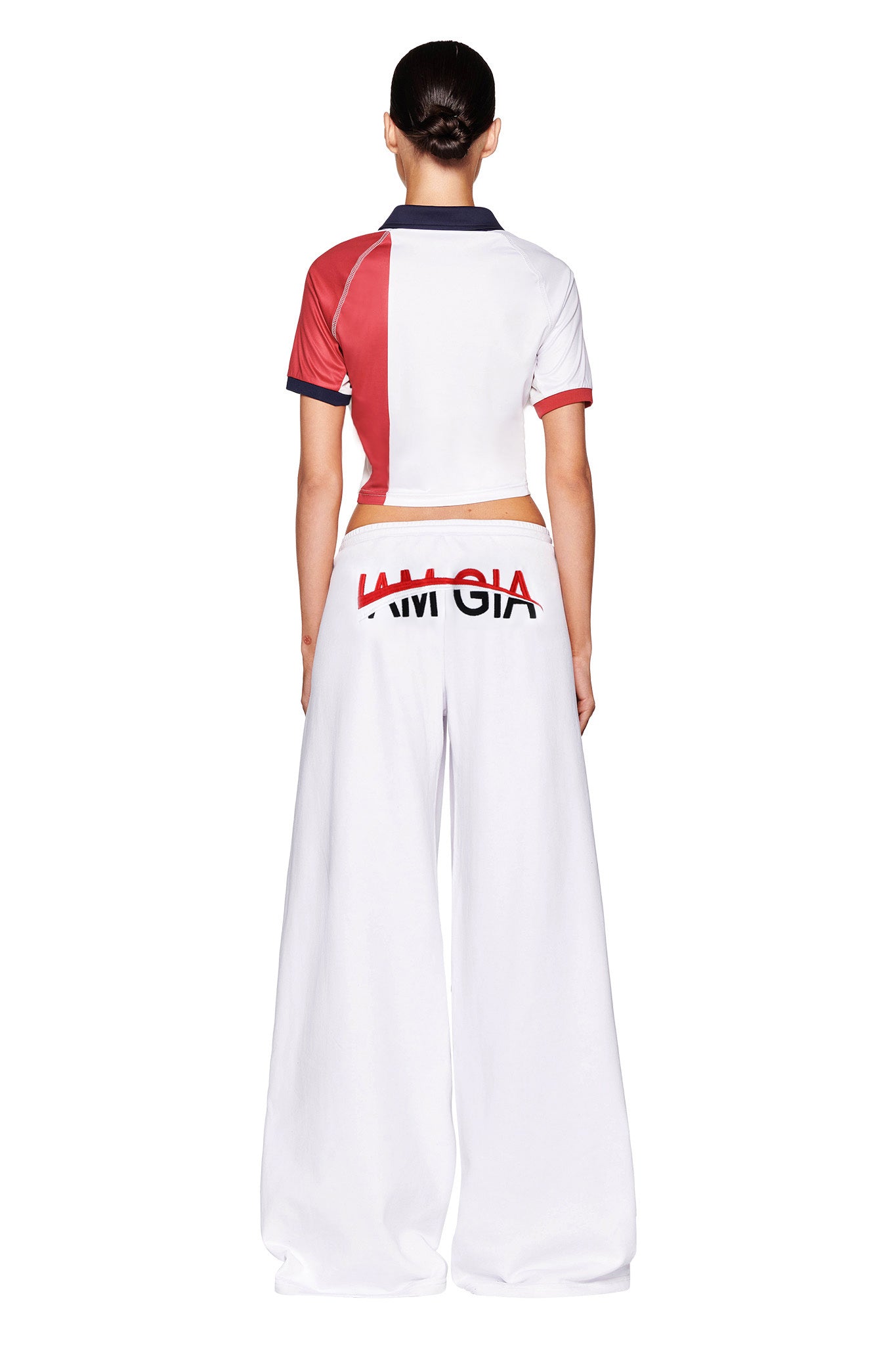 A person with dark hair tied back stands facing away, wearing a short-sleeved, multicolored crop top and the JUSTINE PANT - WHITE by I.AM.GIA, which features large red and black logo lettering near the waistband. The overall style appears sporty.