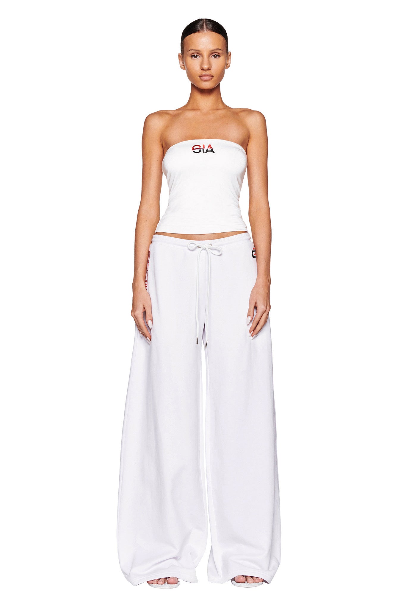 A person stands facing forward against a white background, wearing a white, strapless tube top from I.AM.GIA with an emblem near the neckline, and the JUSTINE PANT - WHITE by I.AM.GIA—extremely wide-leg, high-waisted white pants with a drawstring. Their hair is pulled back neatly.