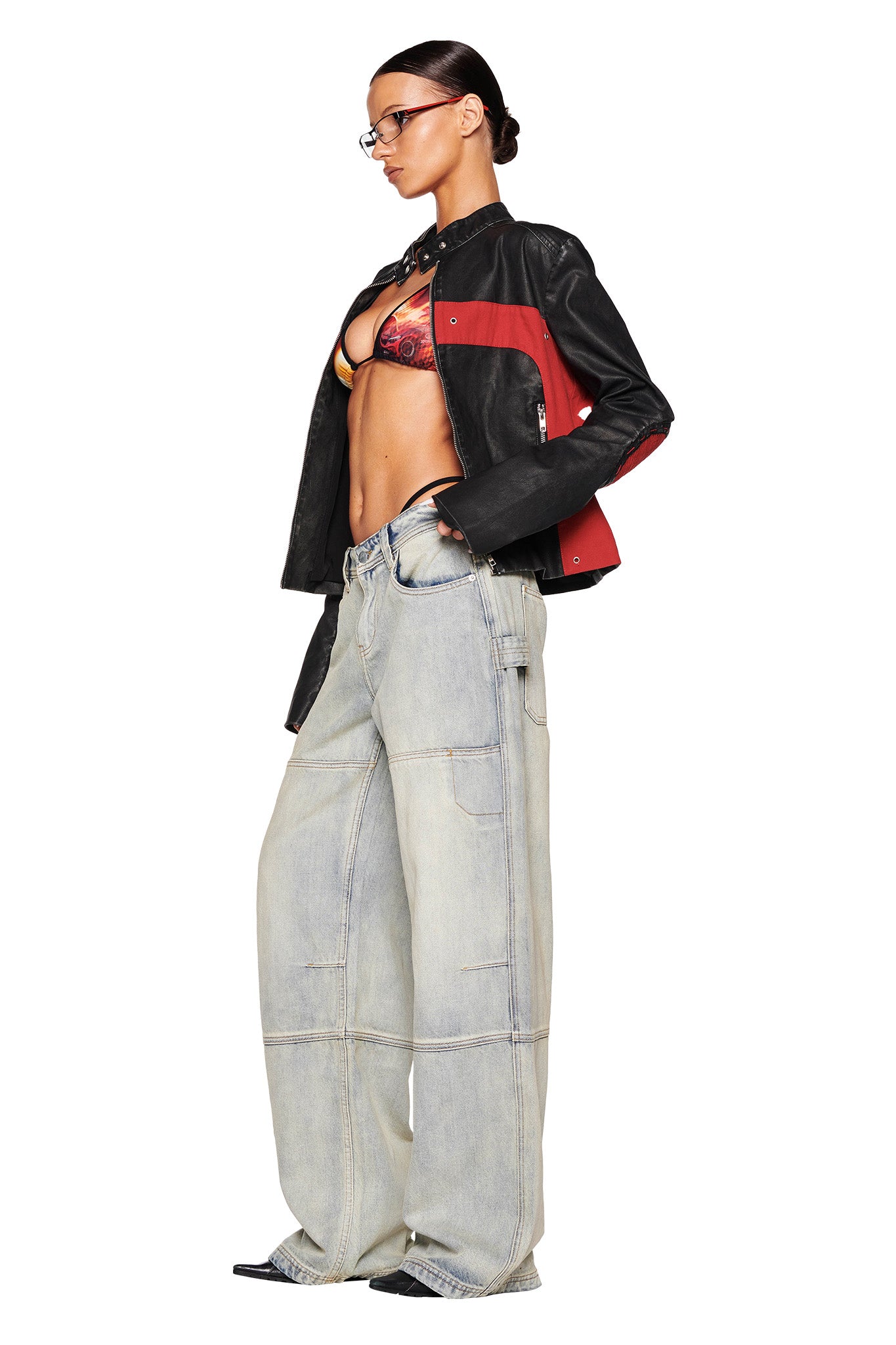 A person stands in profile, sporting an I.AM.GIA black and red motorcycle-style jacket over a patterned bralette and the MISTY JEAN - BLUE : VINTAGE WASH by I.AM.GIA. They have glasses, and their hair is slicked back into a low bun.