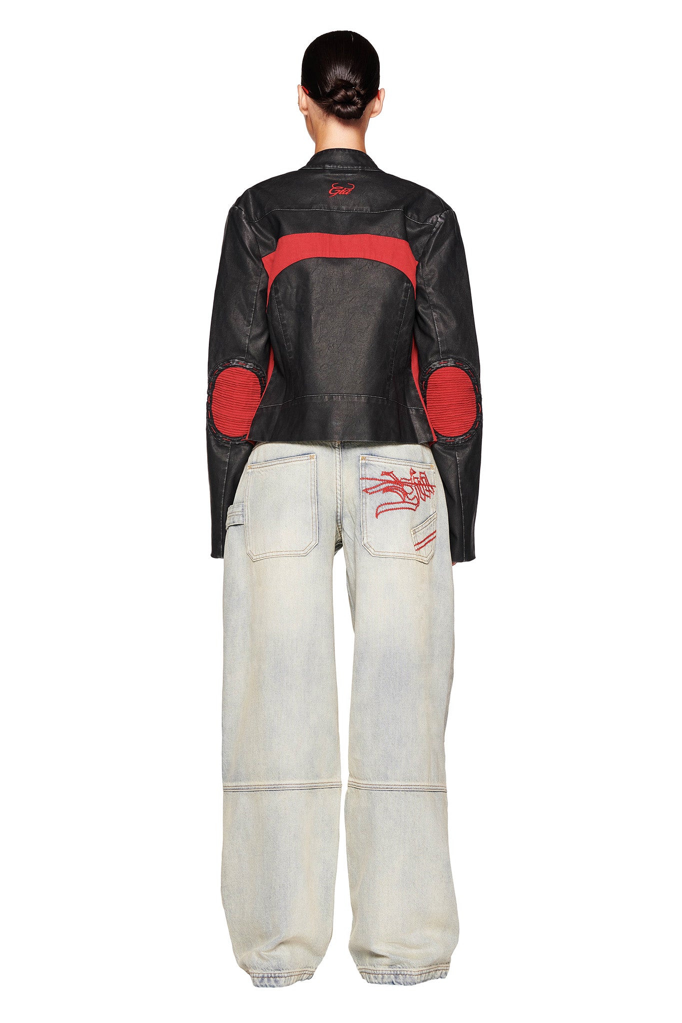 A person is standing with their back to the camera, wearing a black and red leather jacket from I.AM.GIA and light-colored MISTY JEAN - BLUE : VINTAGE WASH baggy jeans featuring red stitching on the back pockets. The hair is styled in a neat, low bun. The pants have elastic cuffs at the ankles.