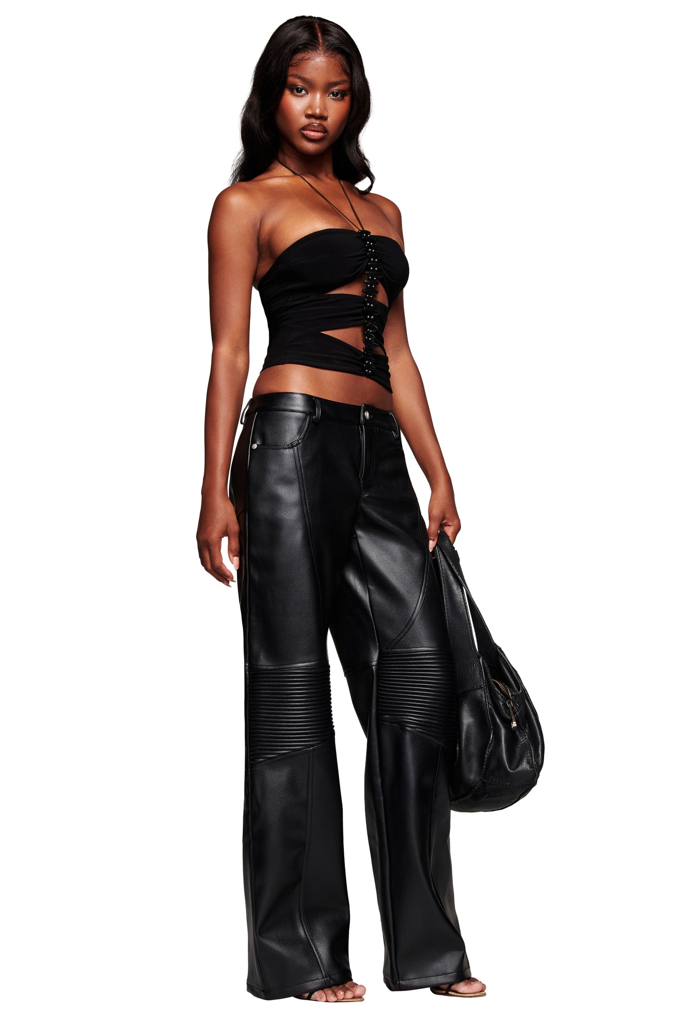 A woman with long, wavy hair confidently models the I.AM.GIA KRISTIE PANT in black, paired with a strapless black crop top and holding a black handbag.