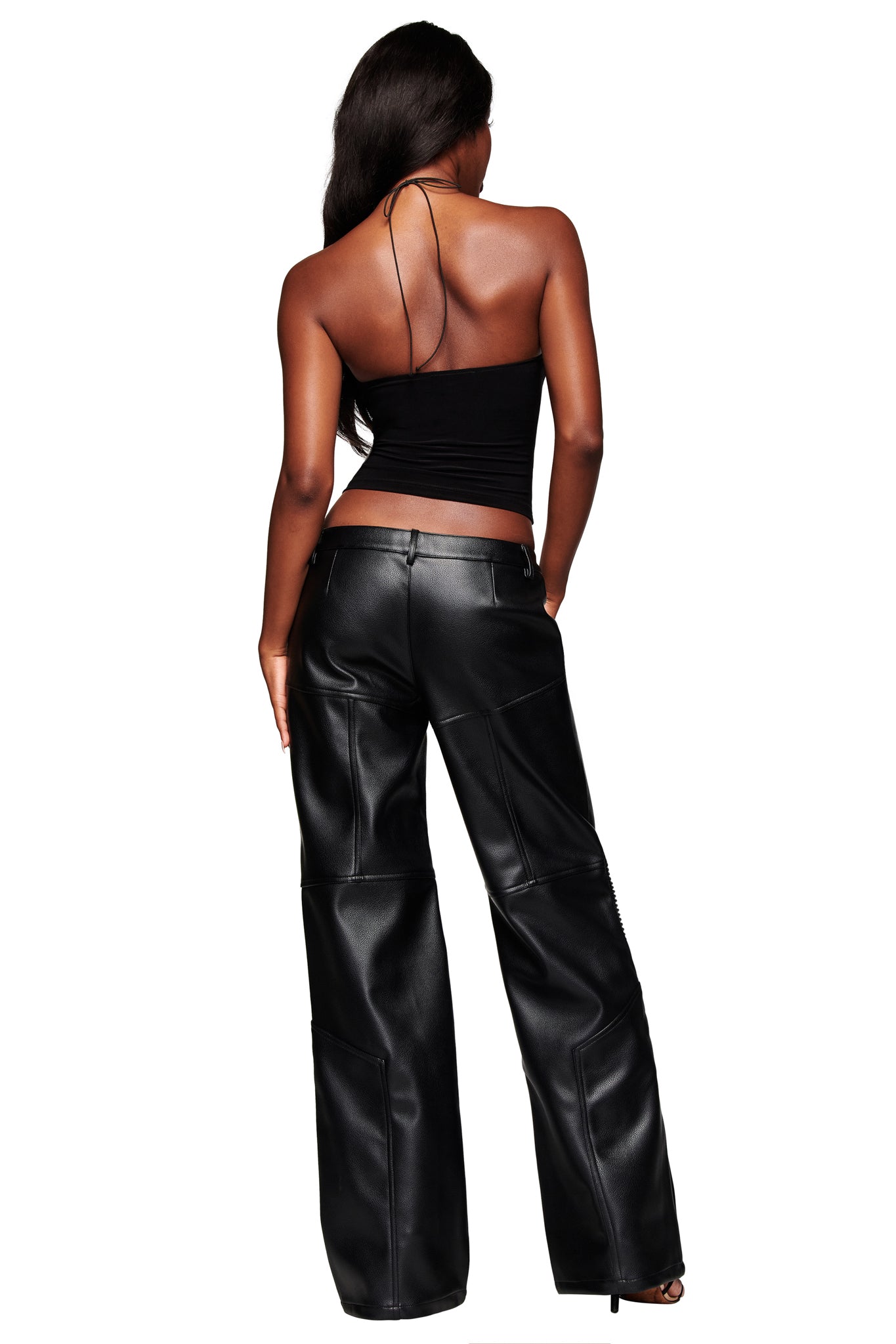 A woman with long dark hair poses against a white background, wearing the KRISTIE PANT - BLACK by I.AM.GIA—low-rise, wide-leg leather pants—paired with a black halter top.