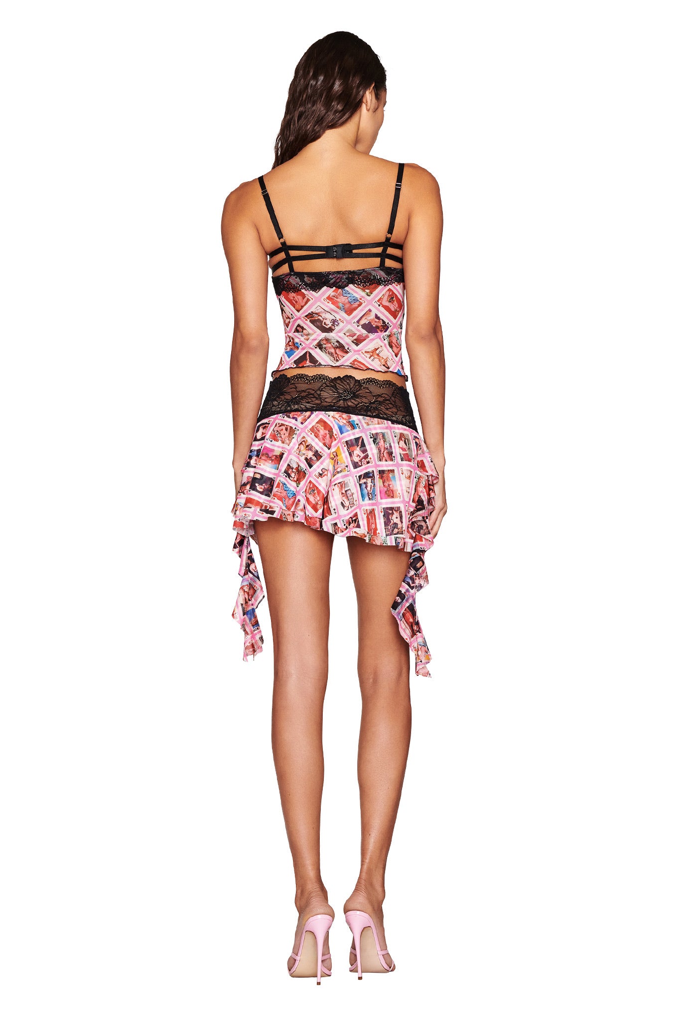 Rear view of a person wearing the I.AM.GIA SILO MINI SKIRT in a colorful pattern. The skirt features black detailing and asymmetrical draping. They are also wearing pink high-heeled shoes.