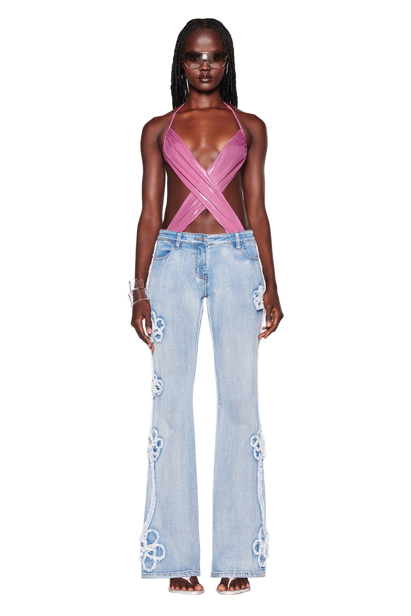 A woman with long braided hair and sunglasses is wearing a pink sleeveless crisscross crop top by I.AM.GIA and the TYLA JEAN - BLUE denim blue high-waisted flare jeans adorned with floral embroidery. She is also accessorized with a chunky bracelet on her right wrist.