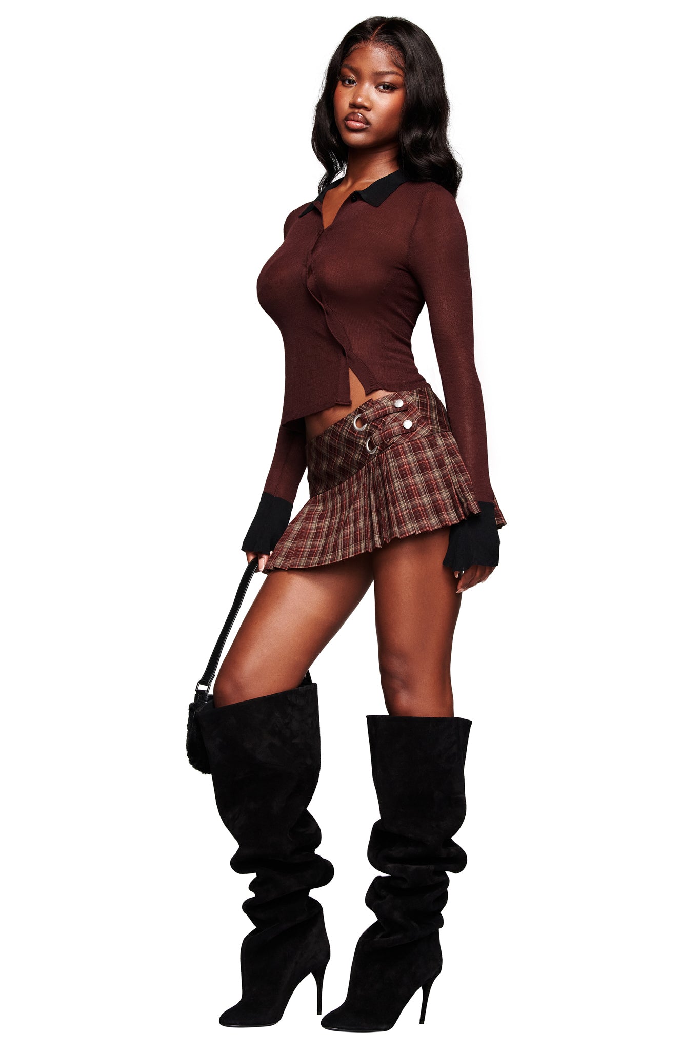 She wears the I.AM.GIA DAVINA TOP in brown, a brown plaid pleated mini skirt with lace-up details, slouchy black knee-high heeled boots, and carries a small black handbag.