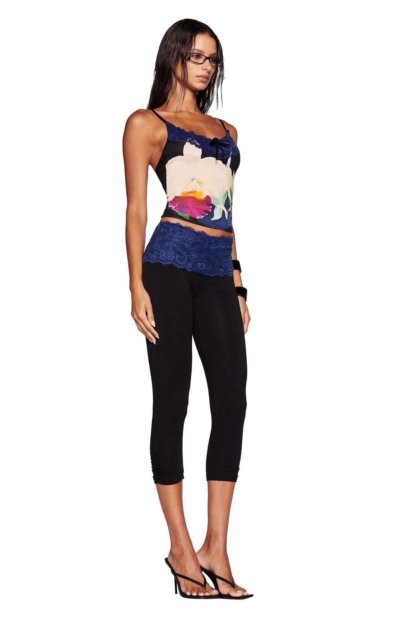 A person with long hair and glasses is wearing the ADELLA TOP in BLACK ORCHID by I.AM.GIA, featuring a custom graphic floral print with blue lace detailing. They are paired with black capri leggings and black high-heeled sandals. The individual stands against a white background, gazing slightly to the side.