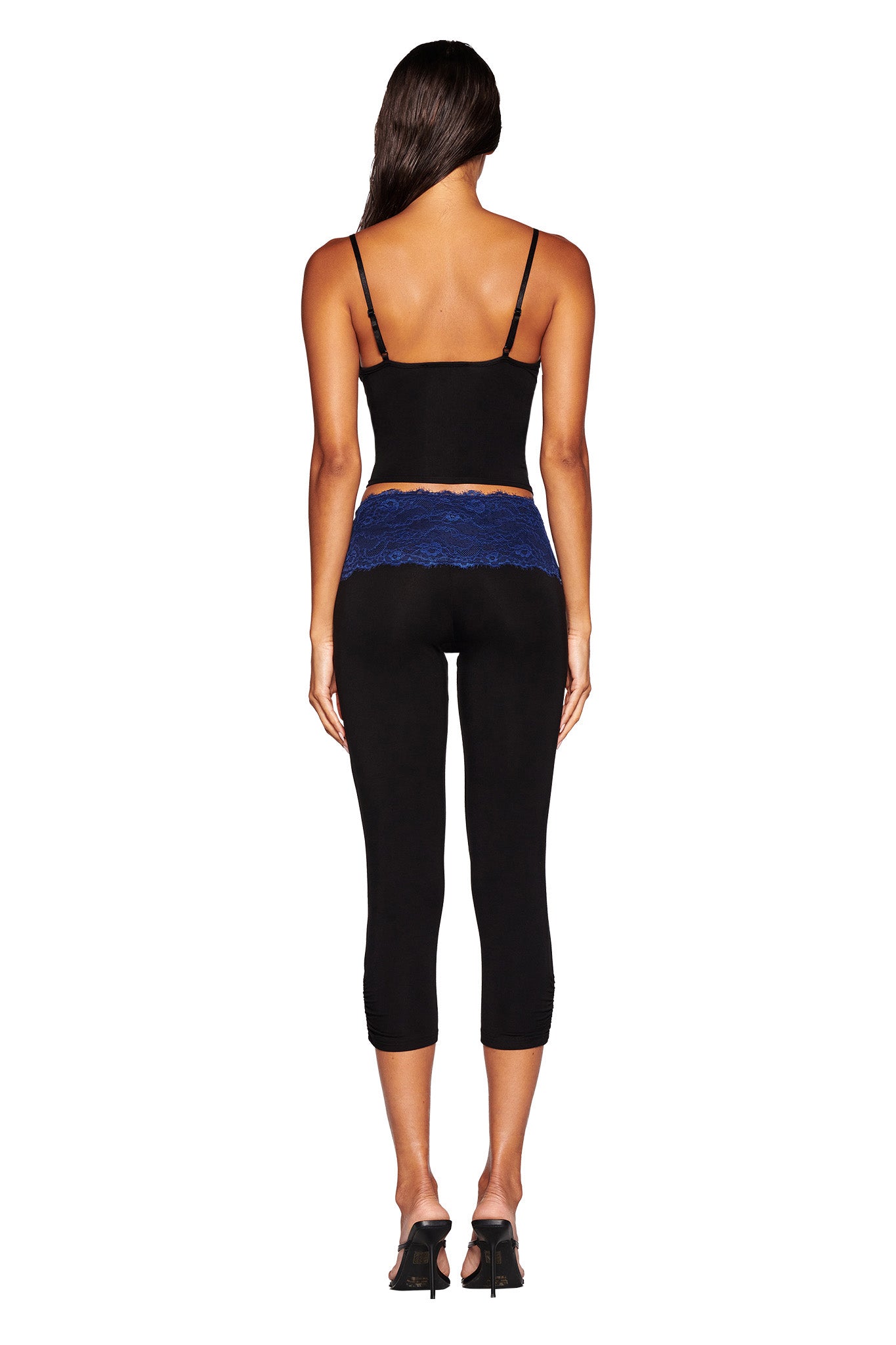 A woman with long brown hair is standing with her back to the camera, showcasing a sleeveless ADELLA TOP - BLACK : FLORAL : BLACK ORCHID from I.AM.GIA. She pairs this top with black cropped leggings featuring a blue lace waistband and black heels. The background is plain white.