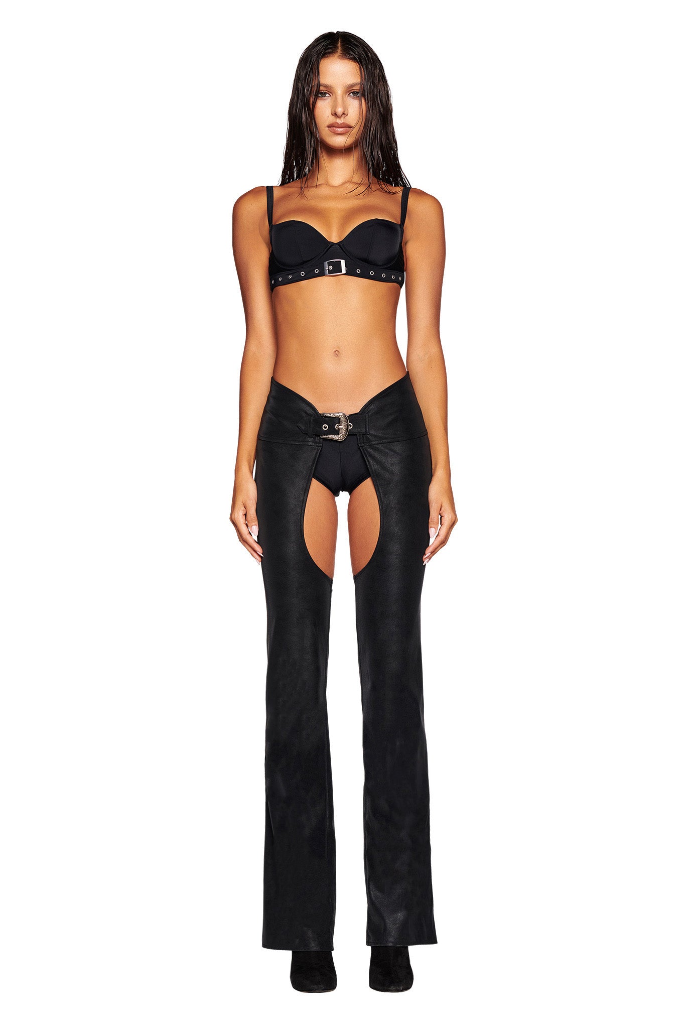 A woman with long wet hair is wearing a black bra and I.AM.GIA's ARIZONA CHAP PANT - BLACK, which features an open thigh design, a flared silhouette, and a mid-rise waist. She pairs the pants with black heeled shoes and stands facing forward, posing against a white background.