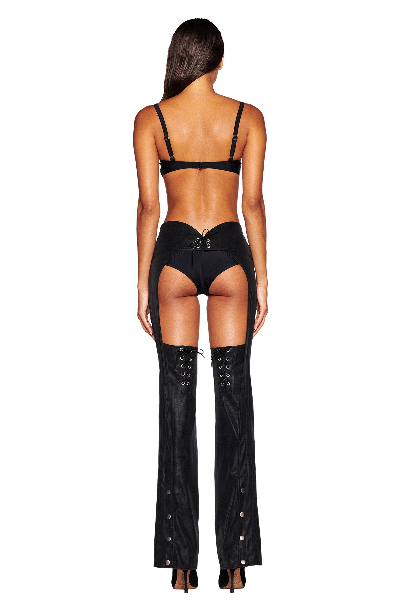 A woman stands confidently with her back to the camera, donning a black lingerie set paired with I.AM.GIA's ARIZONA CHAP PANT in black. The faux leather chap pants feature intricate lace-up details on the back and along the legs, accentuating her mid-rise waist. Her long, straight hair cascades down as she balances effortlessly on high heels.