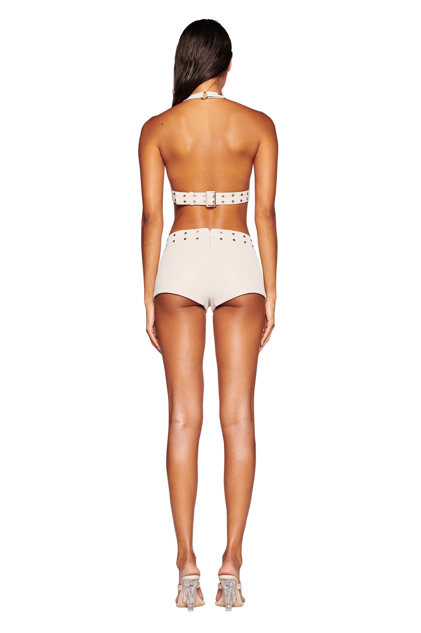 A woman with long, dark hair is shown from the back, wearing the DOLLY TOP - NEUTRAL : TAN by I.AM.GIA, a beige faux leather halter top paired with high-waisted shorts featuring adjustable buckle snap closures. She completes her sleek, crop-length bodycon silhouette with matching high-heeled shoes as she stands against a white background.
