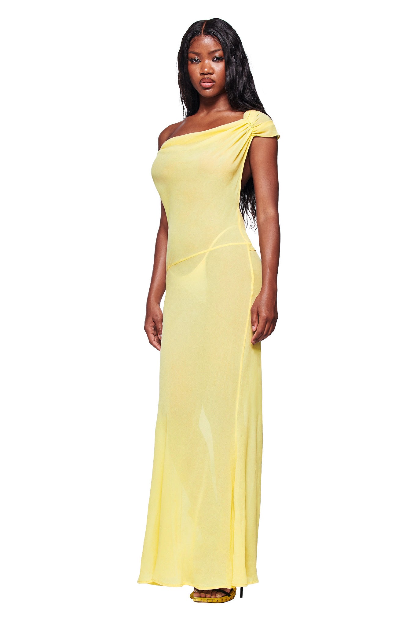 A woman with long, wavy dark hair and a confident expression wears the I.AM.GIA KAHLO MAXI DRESS in yellow—a lightweight georgette off-shoulder maxi with one-shoulder ruching—paired with yellow heels, against a white background.