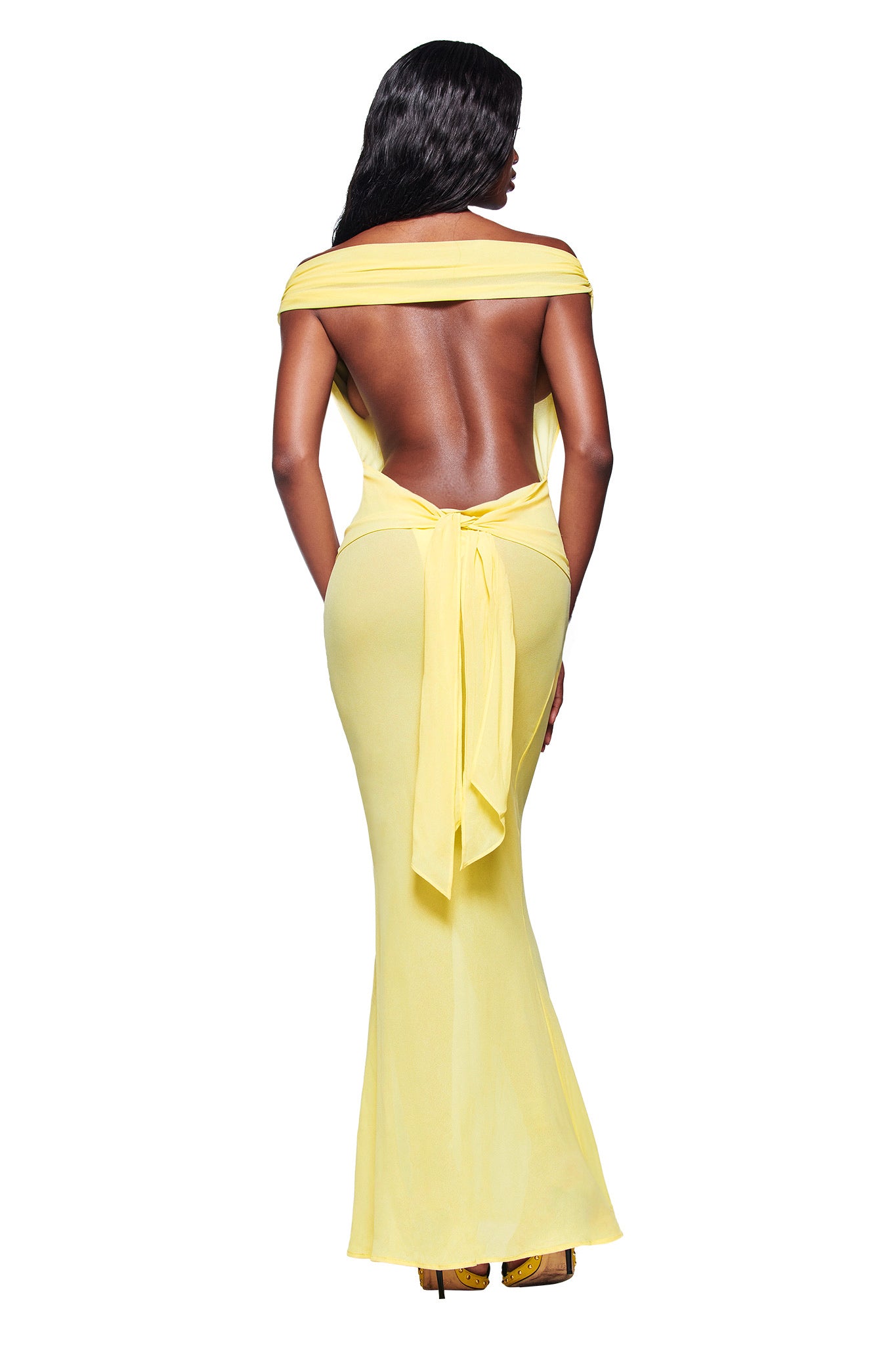 A woman with long dark hair models the I.AM.GIA KAHLO MAXI DRESS in yellow, an off-the-shoulder, backless, floor-length dress with a waist bow, standing against a white background.