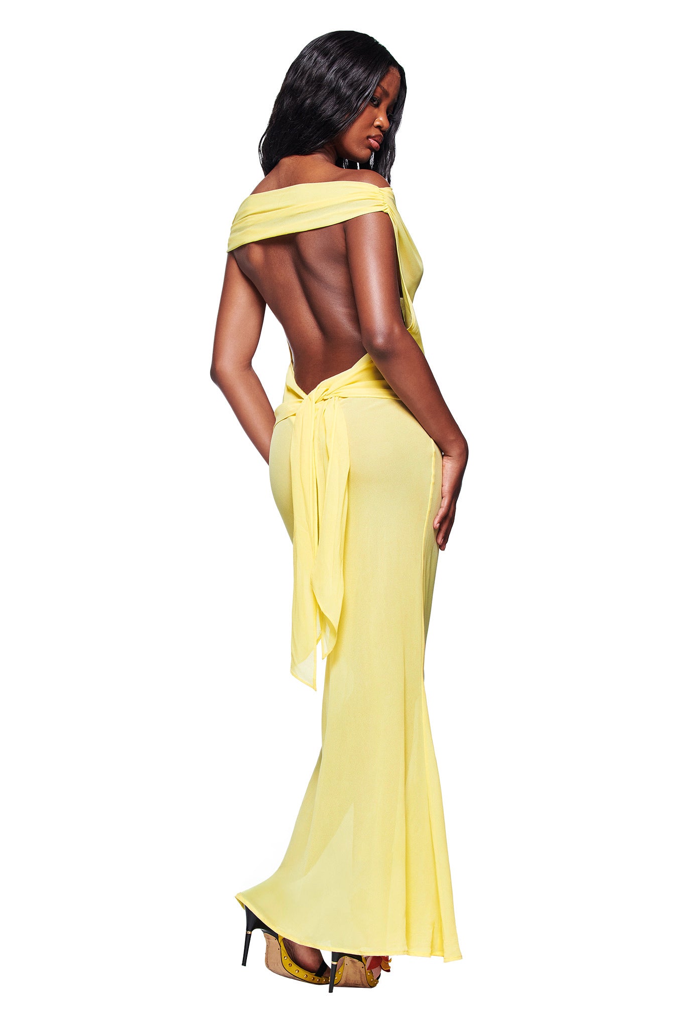 A woman poses confidently in the I.AM.GIA KAHLO MAXI DRESS - YELLOW, a flowing, backless style tied at the lower back. She completes the look with high heels, striking a pose against a white background.