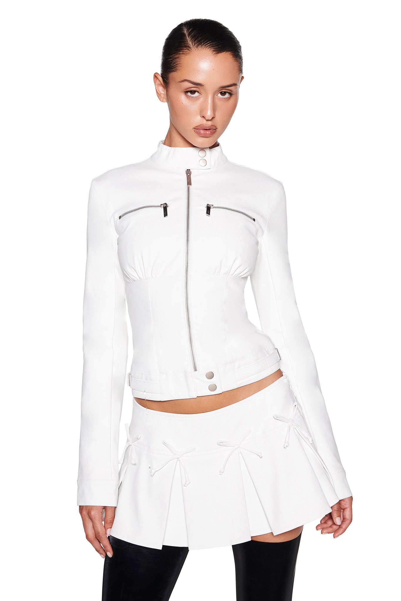 A woman wearing the EVERETTE JACKET - WHITE by I.AM.GIA, showcasing a tailored fit with long sleeves, dual horizontal zippers near the chest, and a high collar. She complements it with a short white skirt adorned with decorative bows and black leggings. Her hair is tied back, and she stands against a plain white background.