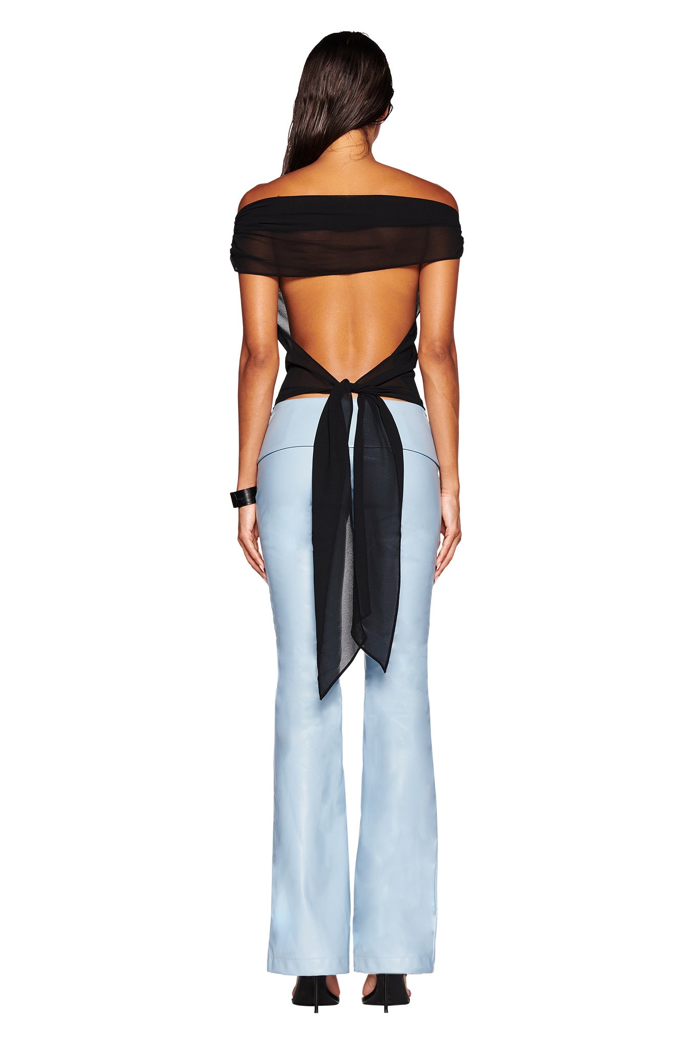 A person stands with their back to the camera, showcasing the I.AM.GIA KAHLO TOP in black. The off-the-shoulder, open-back top is tied with a knot and paired with light blue, high-waisted, flared pants and black heels. The semi-sheer georgette fabric of the top subtly reveals their silhouette as long, dark hair cascades down their back.