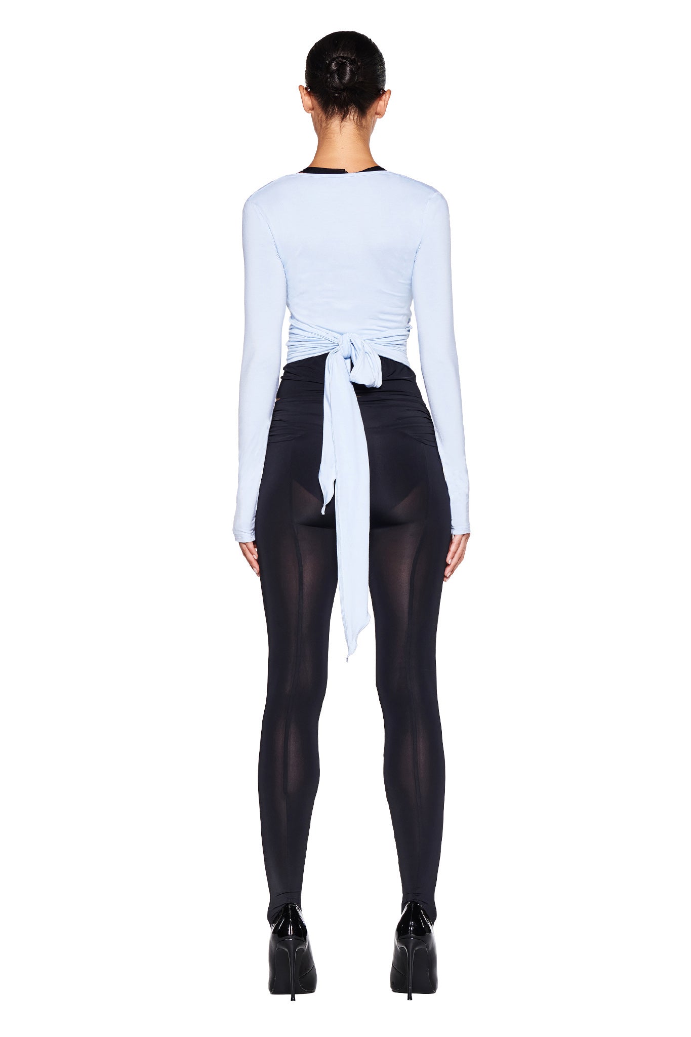 A person stands with their back to the camera, wearing the I.AM.GIA AMRIA TOP in Powder Blue, paired with black sheer leggings and black high-heeled shoes. The person's hair is pulled back.