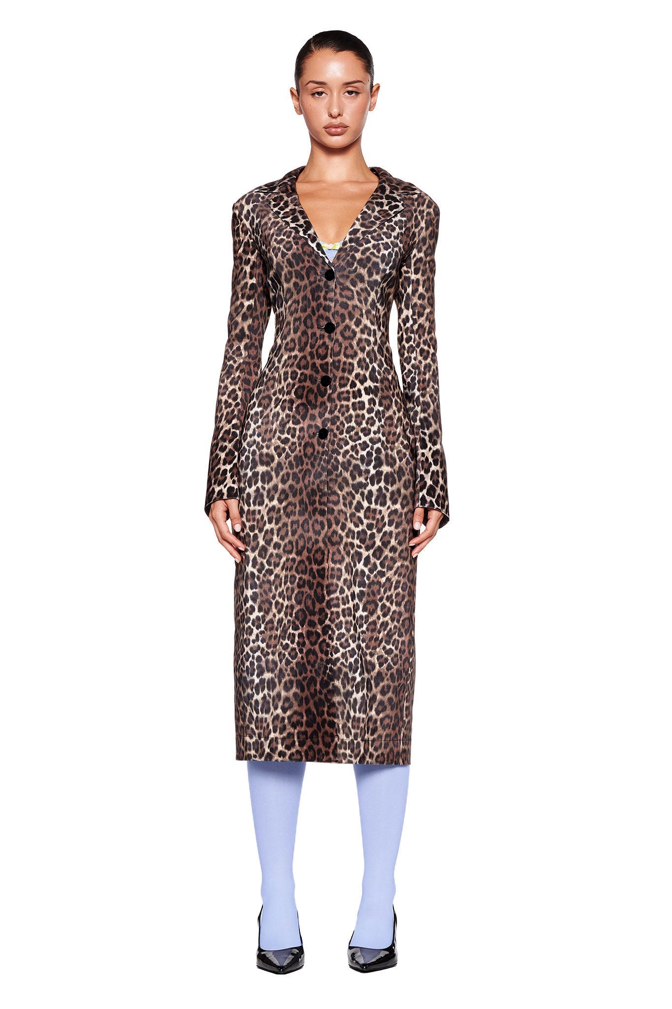 A woman stands facing forward against a white background. She is wearing the UMA COAT - NEUTRAL : LEOPARD from I.AM.GIA, paired with pastel blue tights and black pointed-toe heels. Her hair is pulled back, and she has a neutral expression.