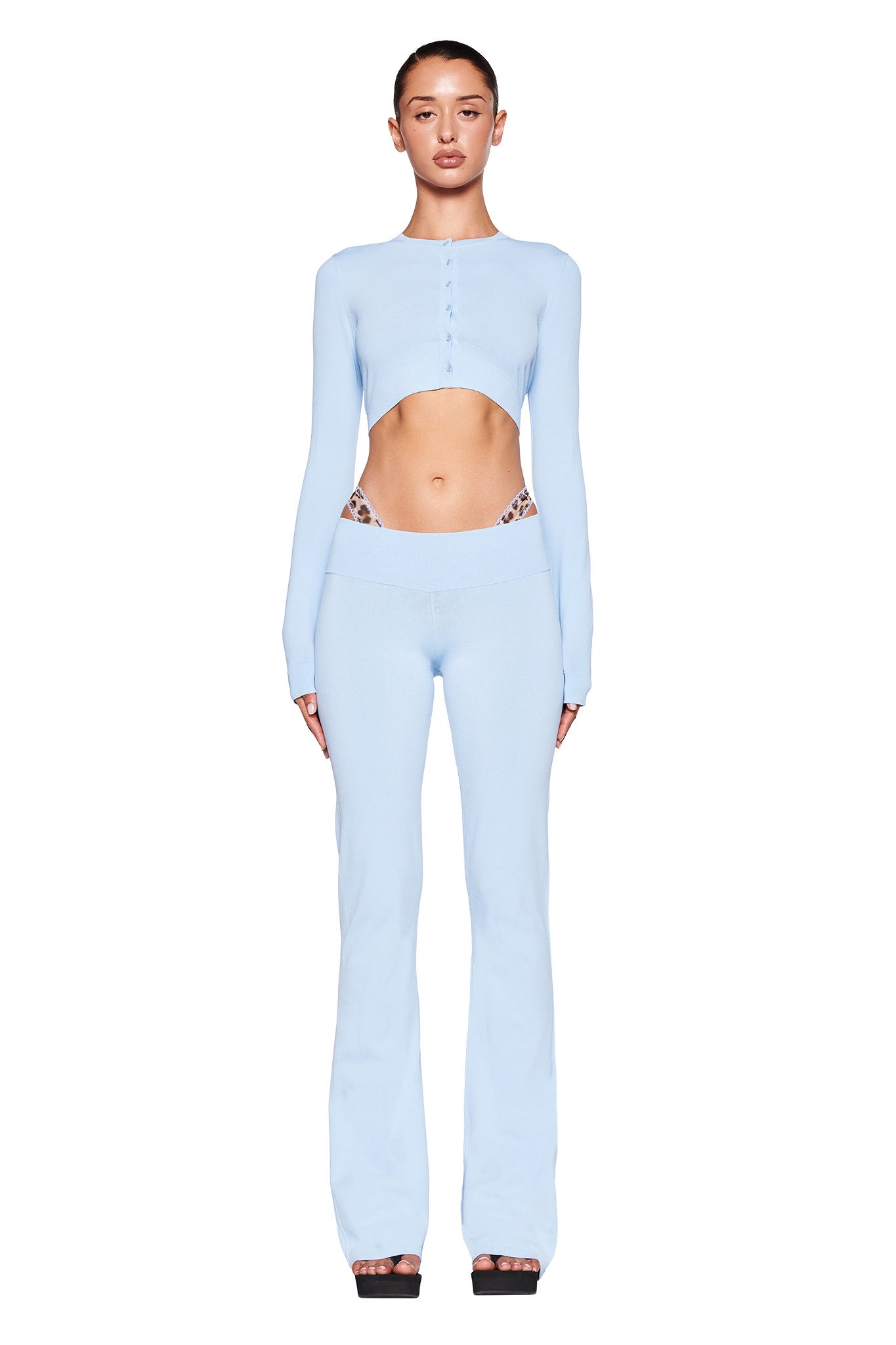 A woman is wearing a light blue, long-sleeved crop top with buttons down the front, paired with the matching AUDRINA PANT - BLUE in powder blue by I.AM.GIA. The outfit reveals her midriff as she stands facing the camera against a plain white background, with her hair pulled back neatly.
