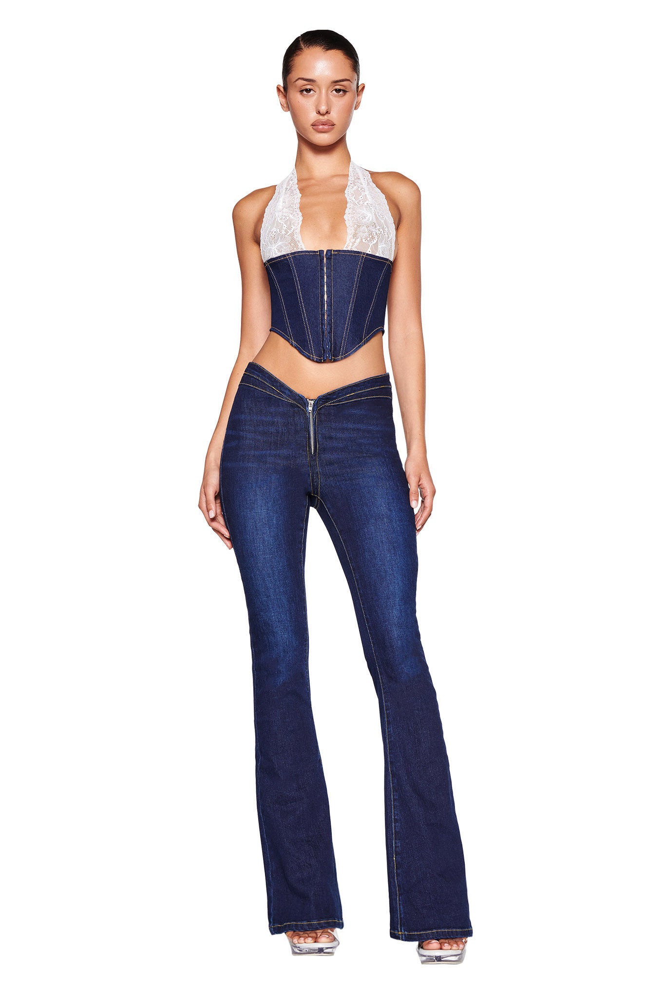 A woman stands against a white background, wearing the ELLENA CORSET - BLUE : DARK WASH by I.AM.GIA, which features a dark blue denim halter corset top with a white lace neckline. She pairs it with dark blue low-rise stretch denim jeans that have zippers at the front and completes her look with clear high-heeled shoes and her hair pulled back.