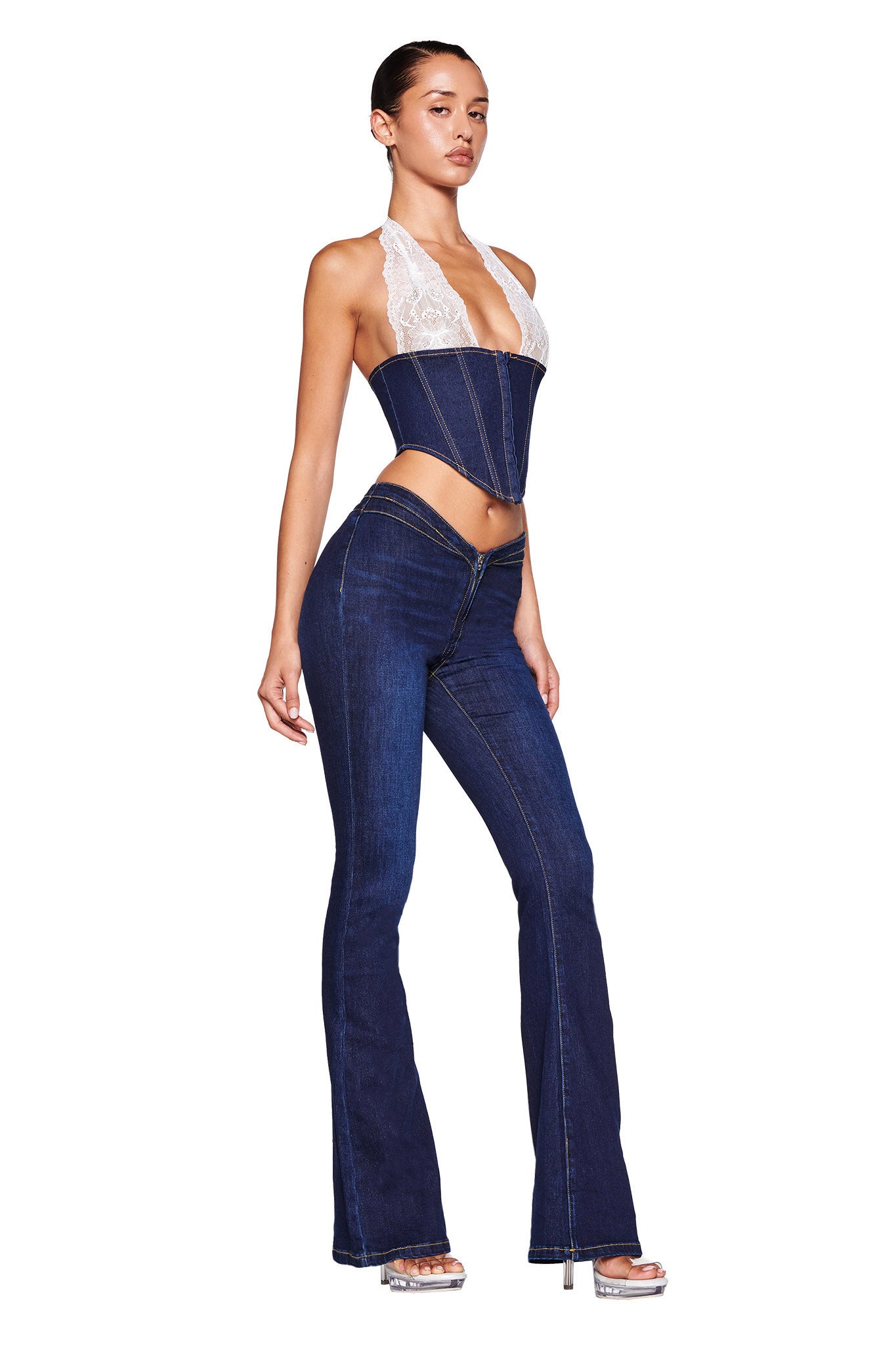 The model is showcasing a chic outfit featuring the ELLENA CORSET - BLUE : DARK WASH from I.AM.GIA, paired with dark blue stretch denim flared pants. The combination has white lace details and offers a bodycon fit that highlights her figure as she poses sideways in clear high-heeled shoes against a white background.