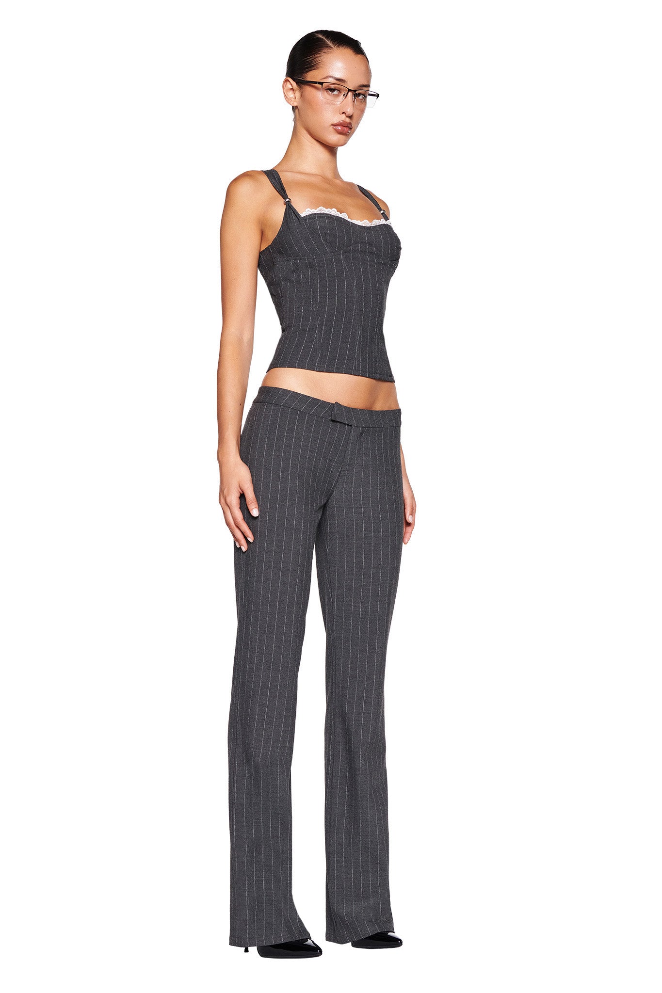 A woman stands against a white background, dressed in a fitted I.AM.GIA MARRION CORSET - GREY : PINSTRIPE and matching pinstripe gray trousers. She wears glasses, black footwear, and has her hair pulled back into a sleek bun, highlighting the tailored silhouette of her ensemble.