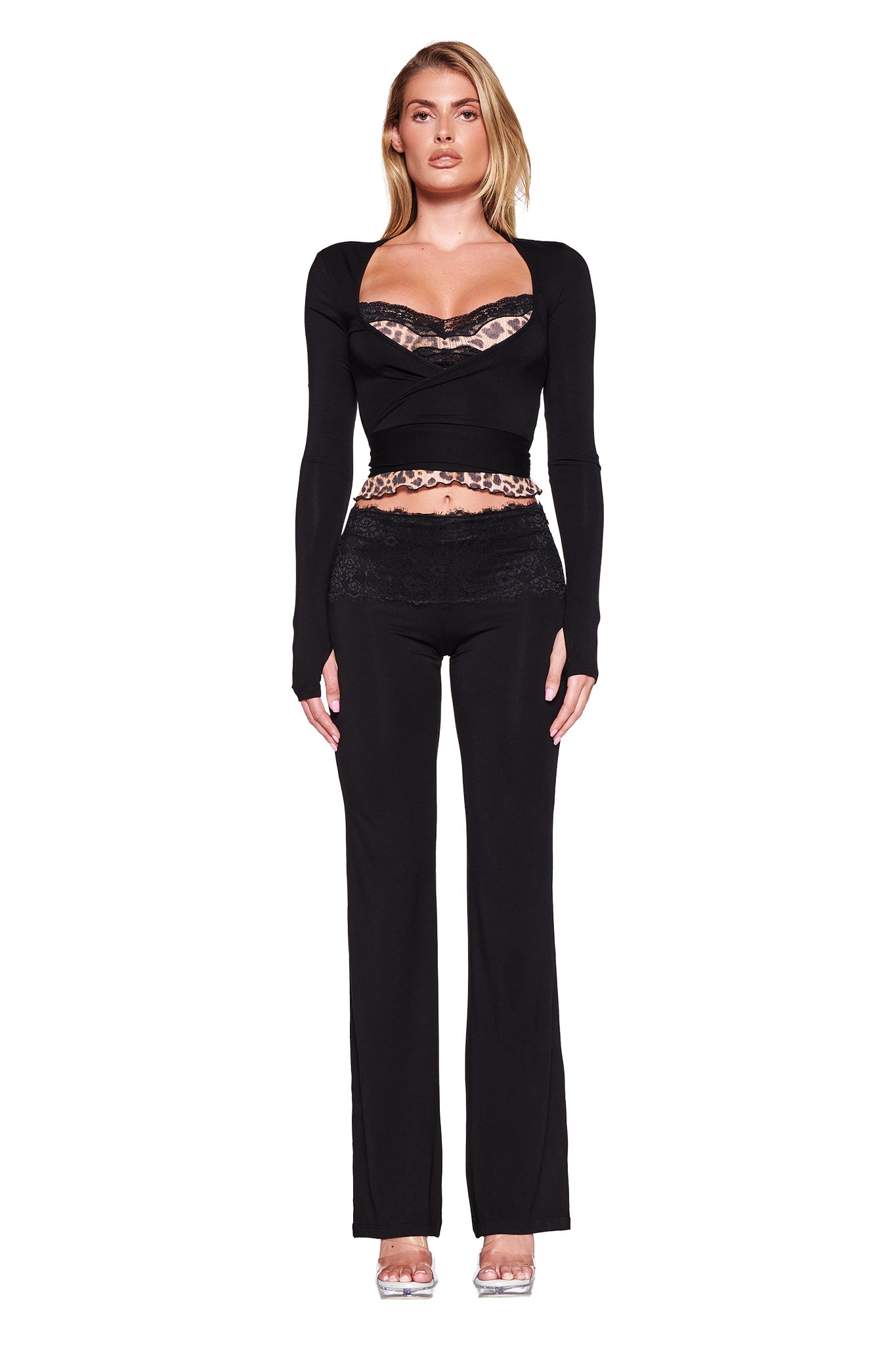 A woman stands against a white background, wearing the I.AM.GIA CASSIA TOP in a neutral leopard print with cut-out details and lace trim, paired with black pants. Her clear high-heeled shoes complement her straight blonde hair cascading over her shoulders. The top features delicate pointelle knits, adding a touch of elegance to the outfit.