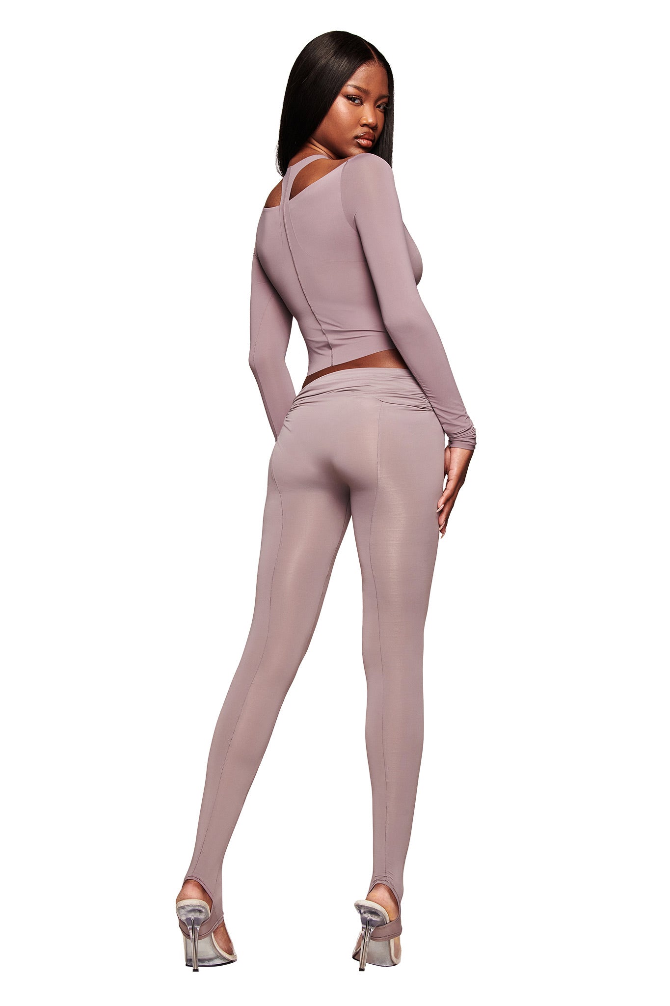 A woman poses against a white background wearing the I.AM.GIA NERINA LONG SLEEVE TOP in dusty lilac with matching high-waisted leggings and silver heels, looking over her shoulder.