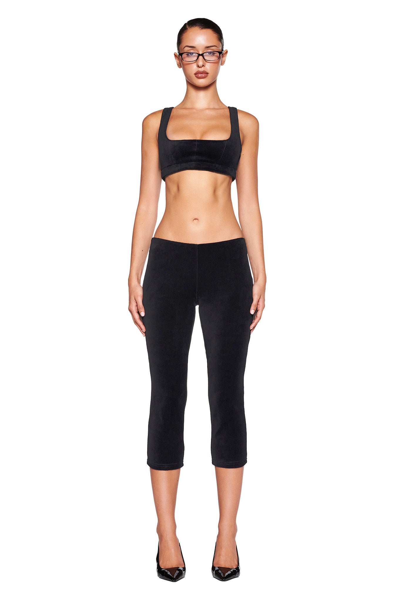 A model stands against a white background wearing a black sports bra paired with the I.AM.GIA LAURA VELVET LEGGING in Black, which features a bodycon fit. She completes her look with black pointed-toe heeled shoes and glasses. Her hair is pulled back, and her expression is neutral.