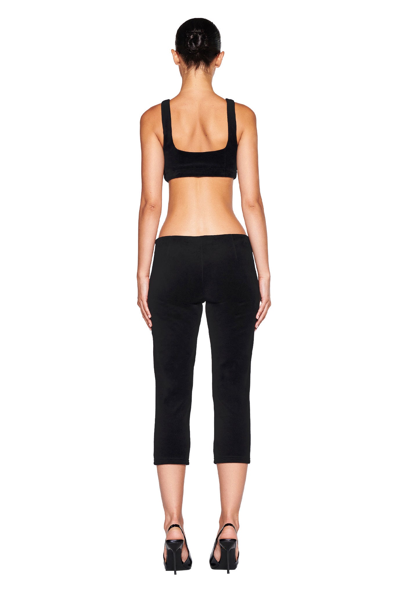 A person with a neat bun hairstyle is seen from the back, wearing a black sports bra, the LAURA VELVET LEGGING - BLACK by I.AM.GIA with a bodycon fit, and black high-heeled sandals. The background is plain white.