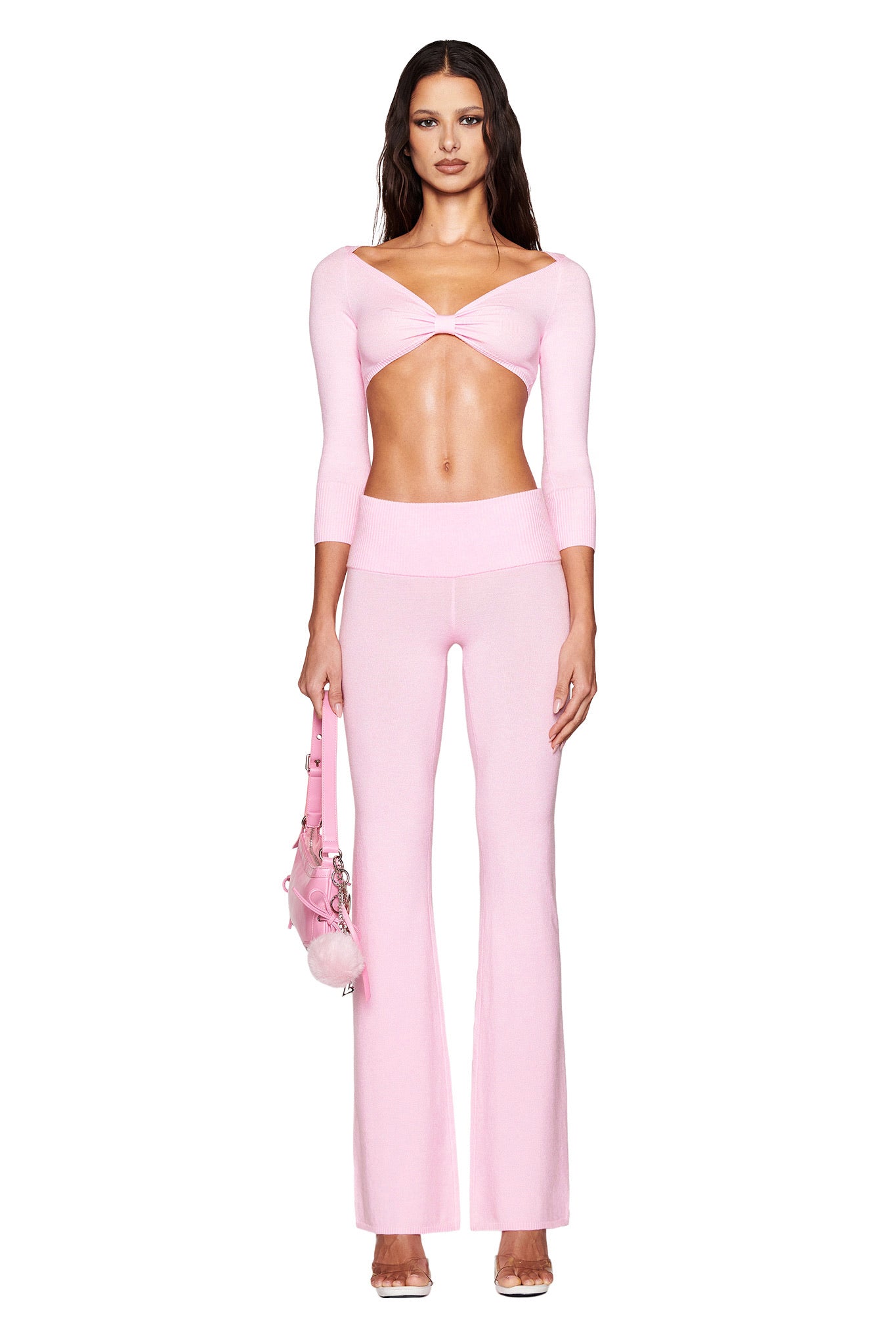 A woman wearing the I.AM.GIA MORGANA PANT in baby pink poses against a white backdrop. Her elegant outfit includes a knit cropped top with three-quarter sleeves and high-waisted flare pants. She complements her look with a pom-pom adorned pink bag and clear high-heeled shoes.
