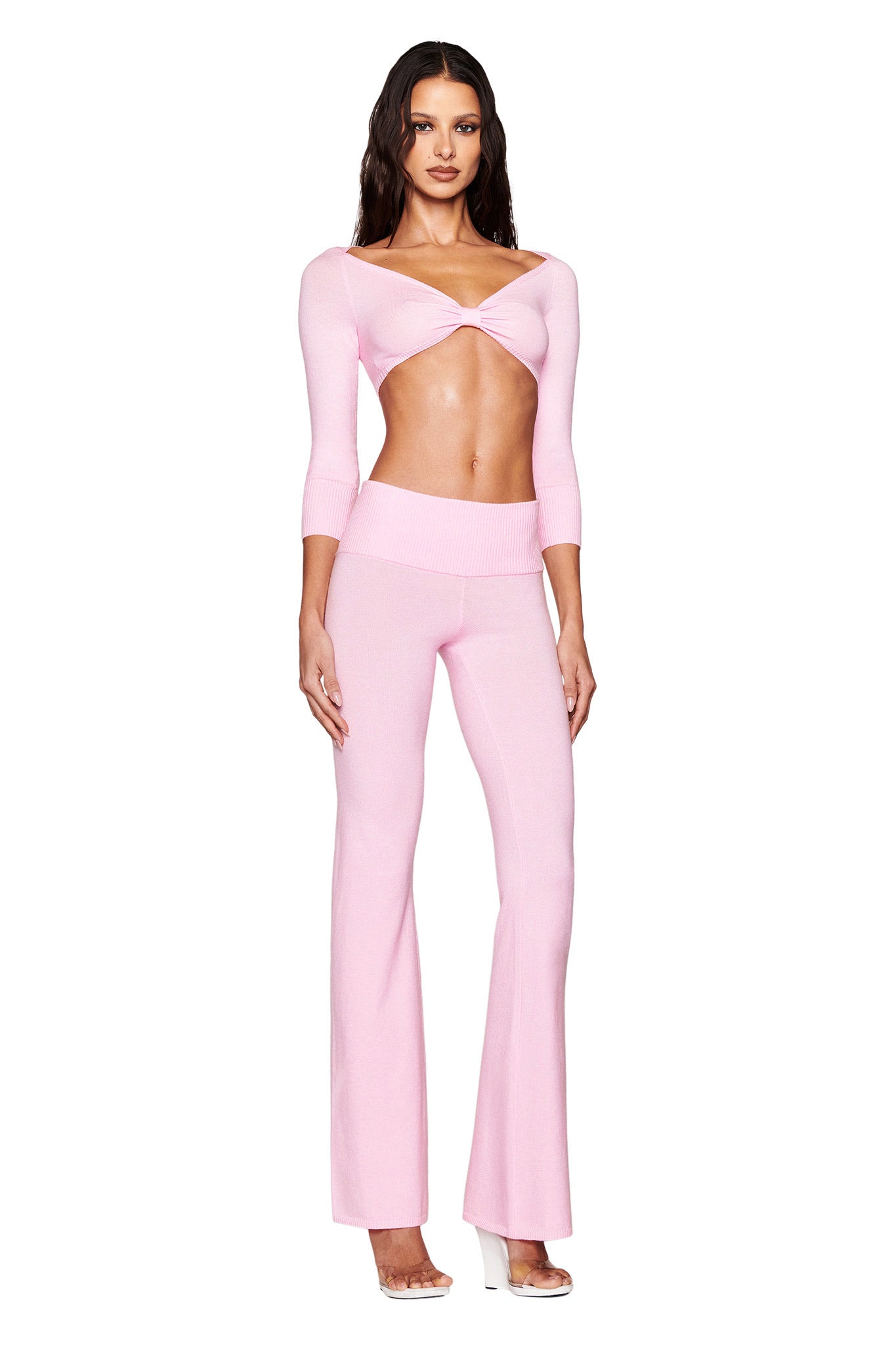 A person models the MORGANA CROP TOP - PINK by I.AM.GIA, showcasing a minimalist super crop with twisted ultra-soft knit, three-quarter sleeves, and high-waisted flared pants. The bodycon fit pairs well with clear high heels against a white background.