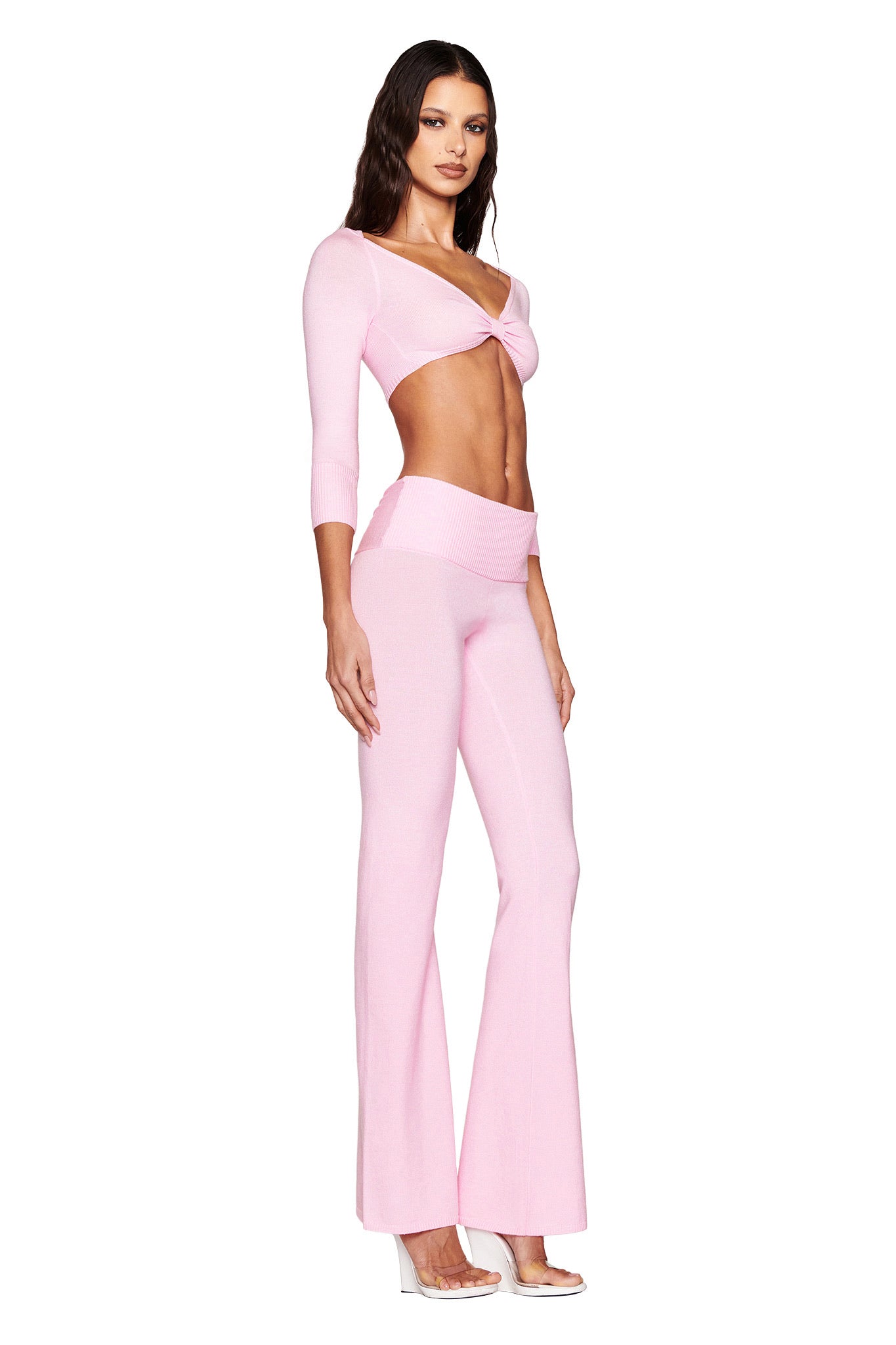 A woman with long, dark hair poses in a baby pink outfit from I.AM.GIA against a white backdrop, featuring a long-sleeve twisted crop top and the MORGANA PANT - PINK, high-waisted and flared in chic knit. She pairs this look with clear high heels.