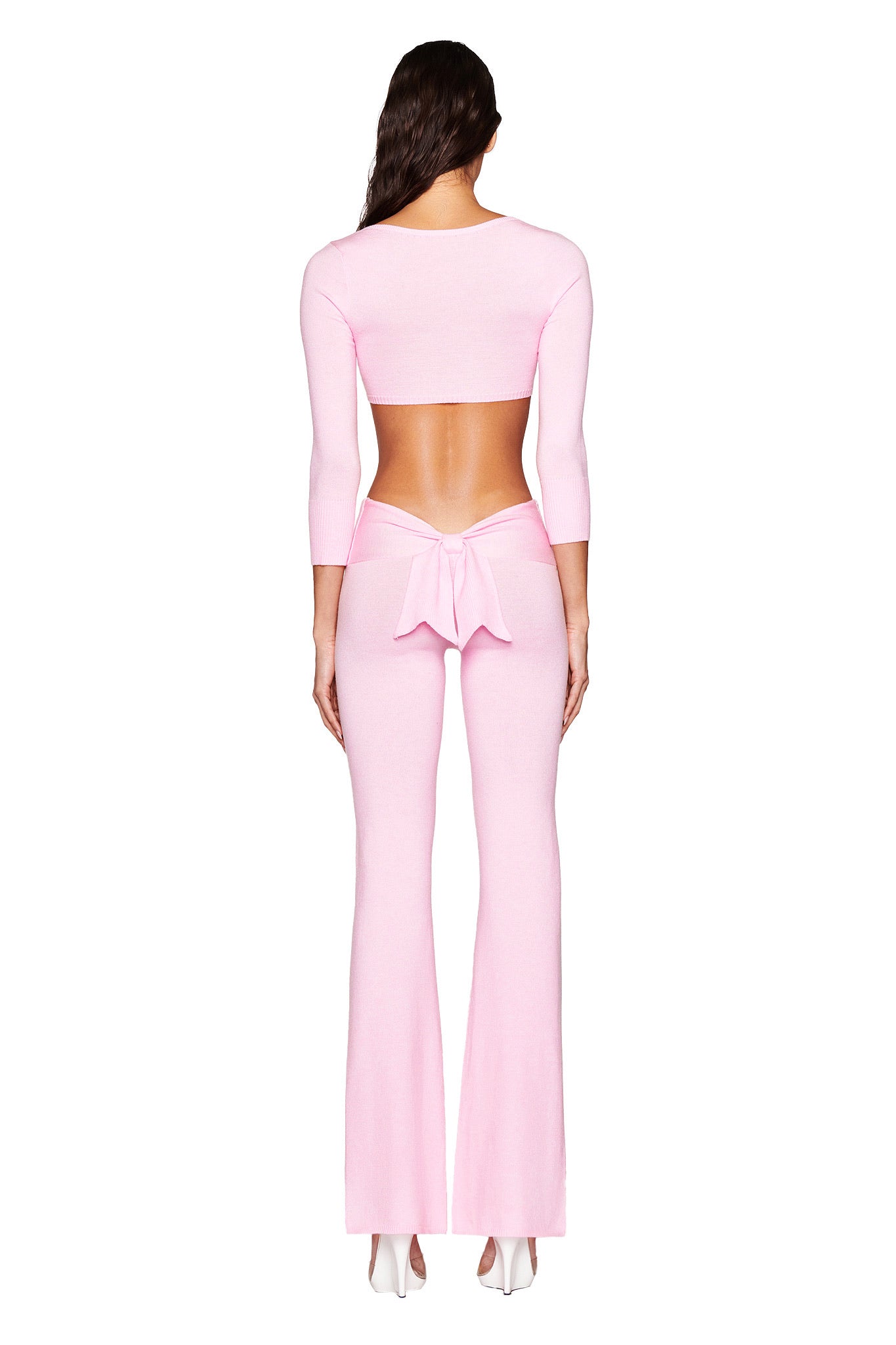 A person stands facing away, dressed in the MORGANA CROP TOP by I.AM.GIA—a light pink, backless bodycon fit with long sleeves and a bow at the lower back. White shoes complement the look, while long dark hair adds elegance to this ultra-soft knit ensemble.