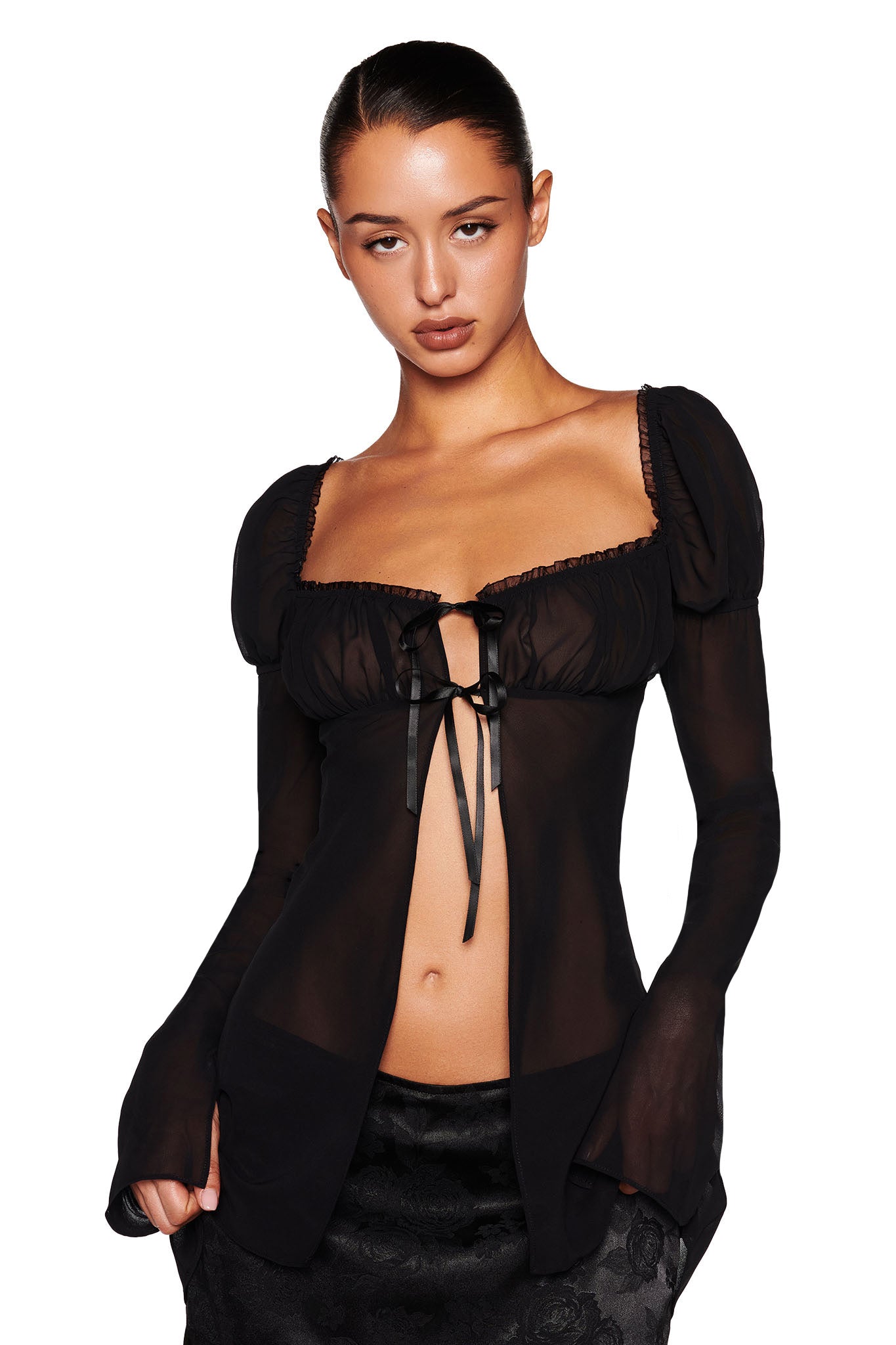 A person wearing the LINDSAY TOP in black by I.AM.GIA, featuring sheer fabric with puffed shoulders, a cinched bust, and a ribbon tie, paired beautifully with a floral-patterned skirt. Their hair is pulled back, highlighting a neutral expression against the plain white background.