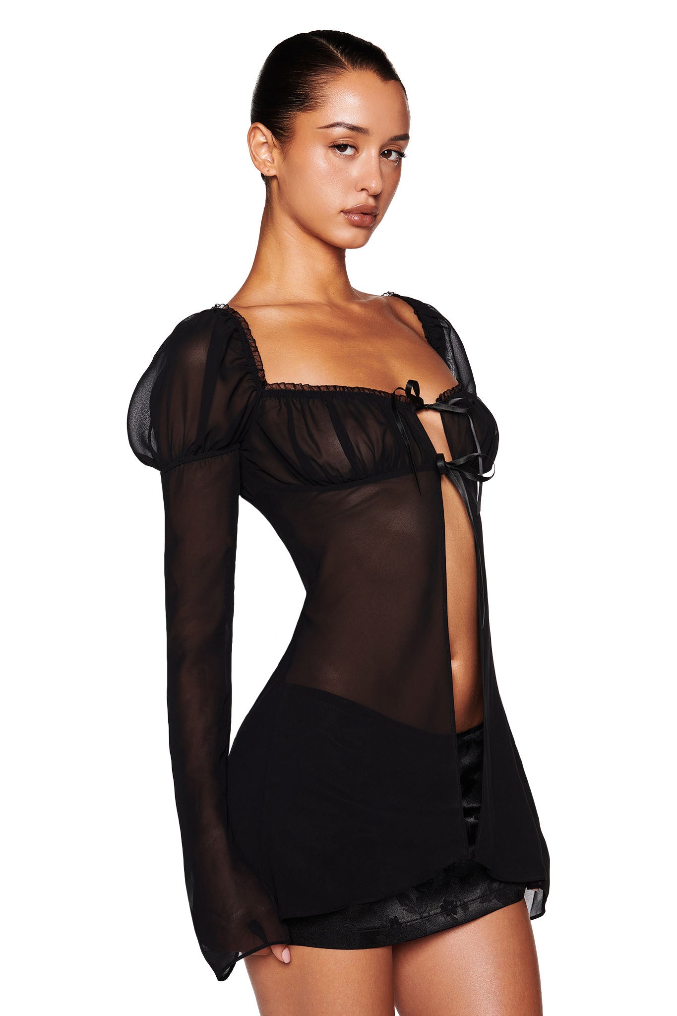 A person with hair pulled back is wearing the LINDSAY TOP by I.AM.GIA, a black sheer top featuring puffed shoulders and a bow at the neckline. They are also wearing a short black skirt against a plain white background.