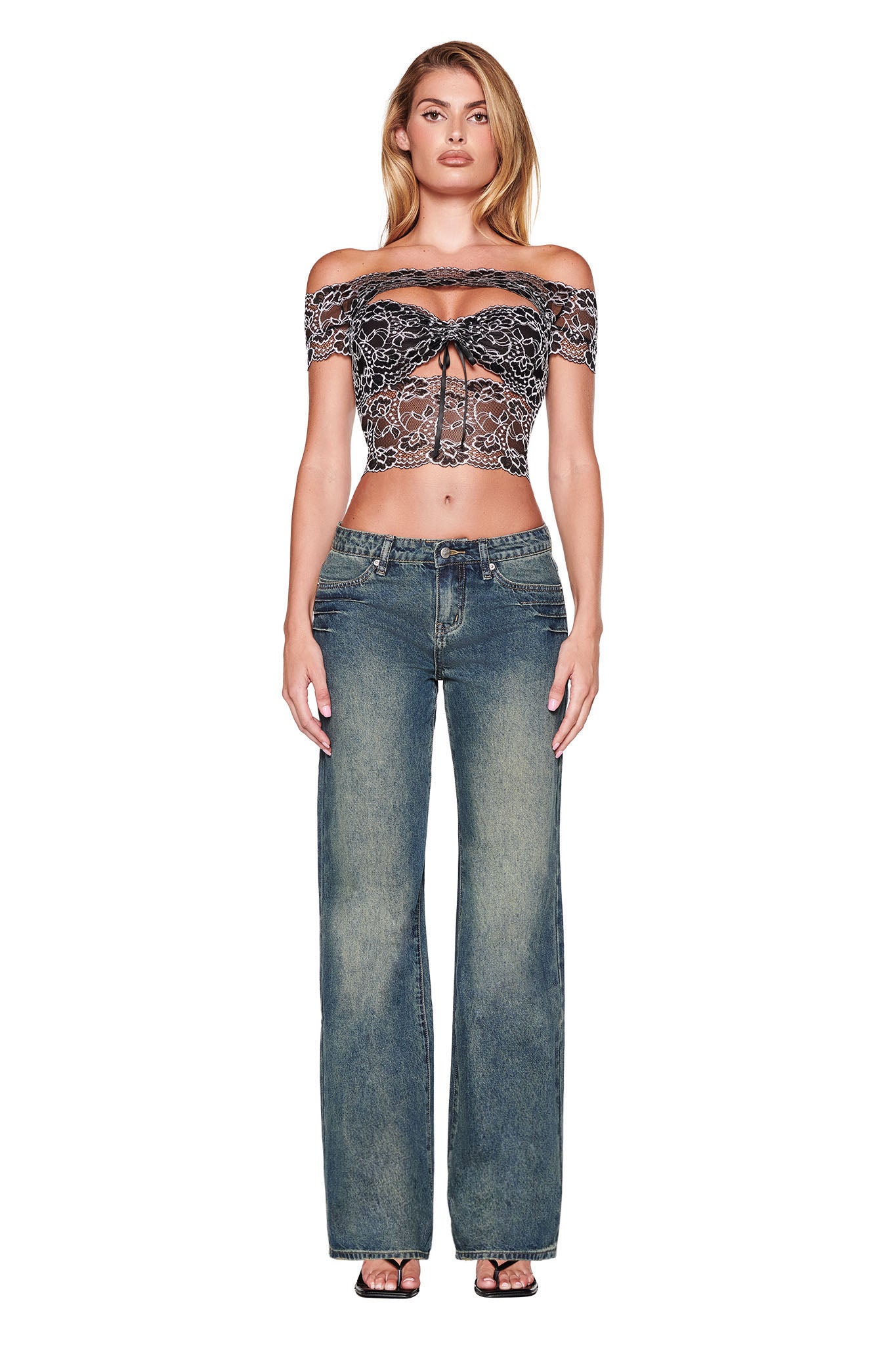 A person stands against a white background wearing a black off-the-shoulder crop top with lace detailing and the I.AM.GIA DYLAN JEAN in blue denim. They are also wearing black sandals and have long hair down, effortlessly exuding chic style in their wide-leg fit.