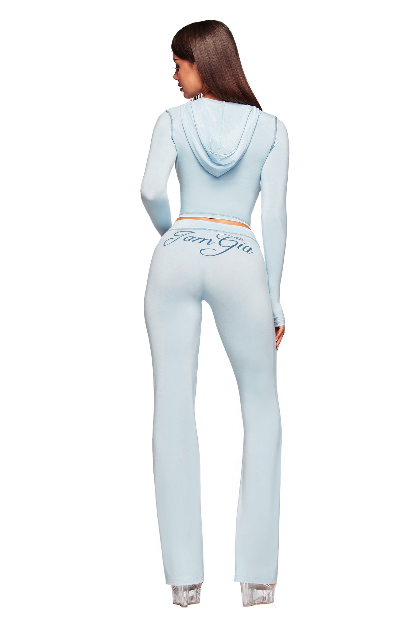A woman is seen from the back wearing the I.AM.GIA BLARE HOODIE in baby blue with matching bodycon flared pants featuring "IamGia" script across the rear, paired with clear high-heeled shoes.